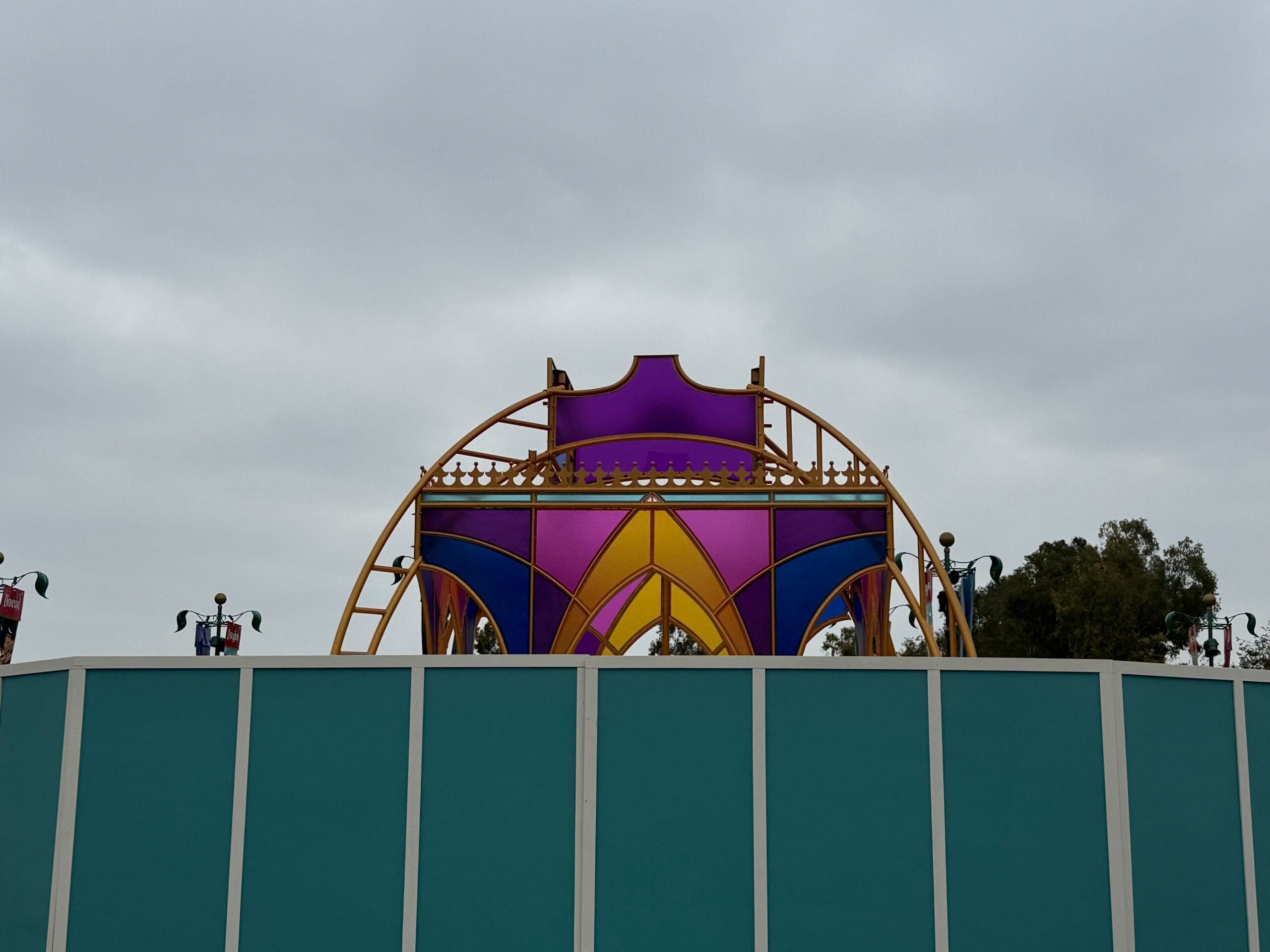 A colorful 70th Anniversary Castle Sculpture rises above a teal construction wall under a cloudy sky at Disneyland Resort.
