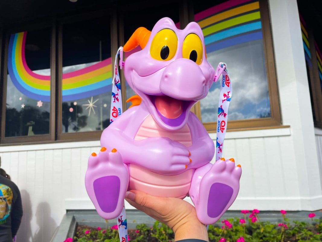 A hand holds a Disney Popcorn Bucket shaped like a purple dragon in front of rainbow-decorated windows.