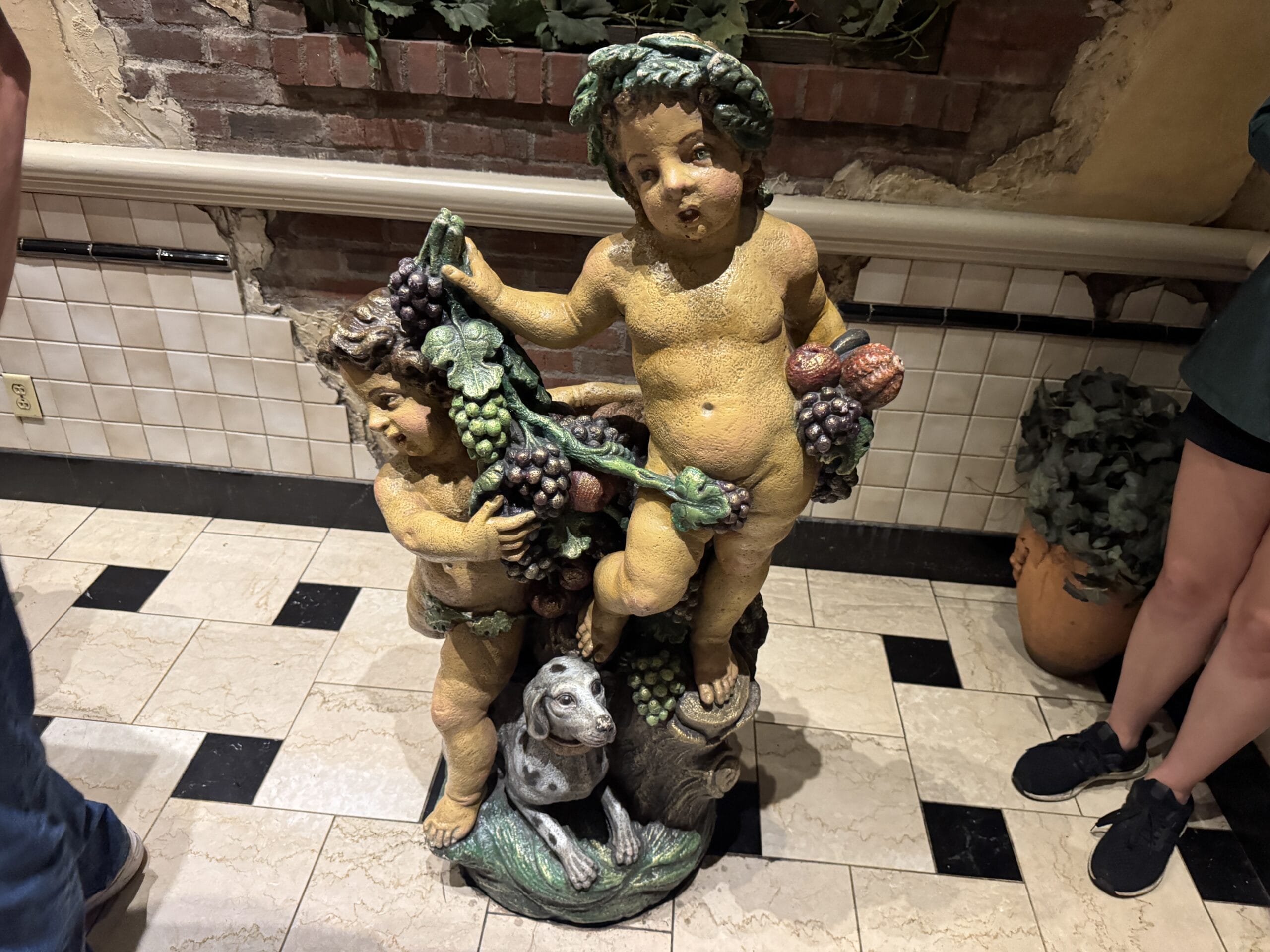 A statue of two cherubs with fruit and a dog stands on tiled floor at Mama Melrose’s Ristorante Italiano, perfect for photos.