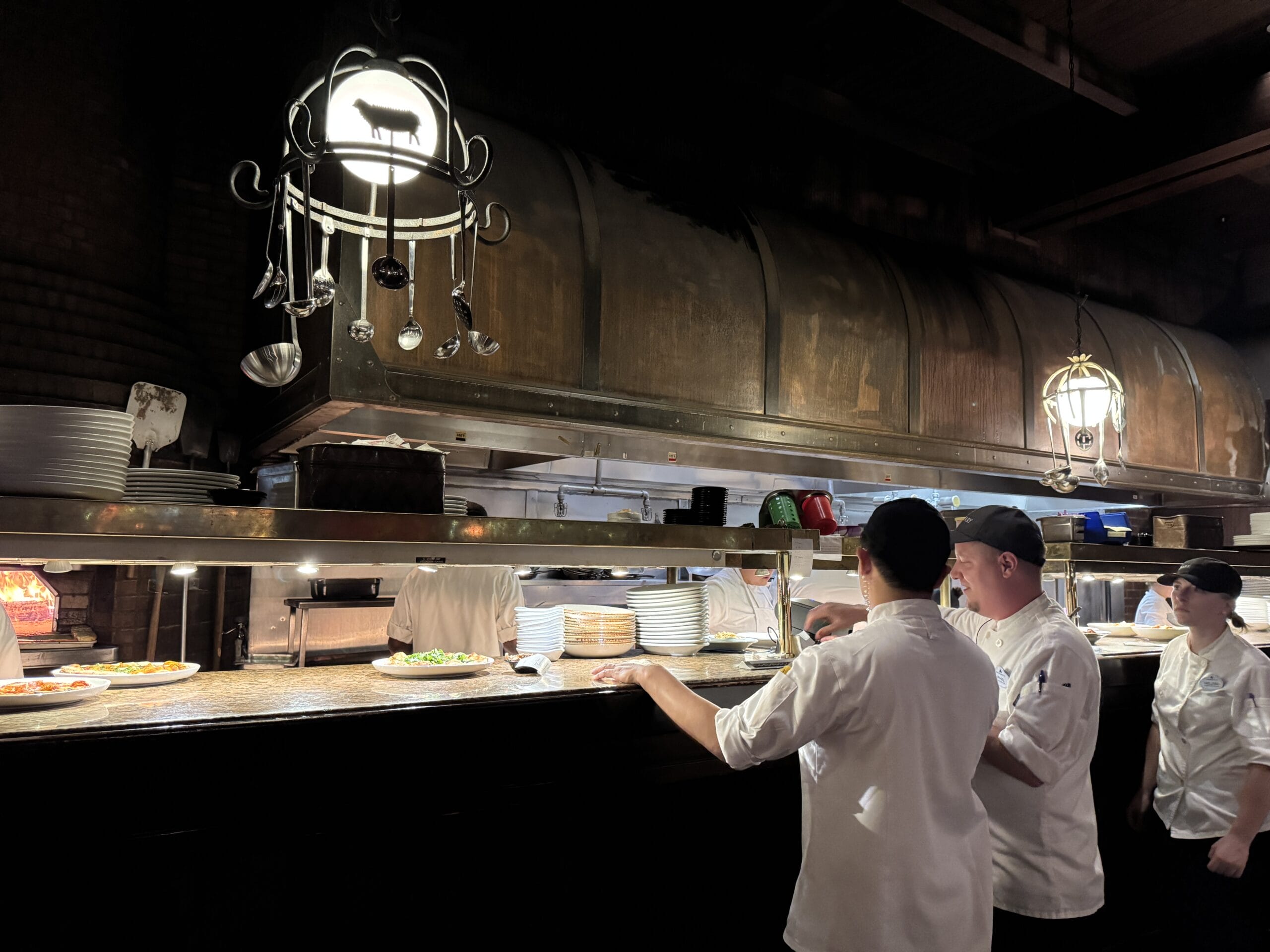 Chefs in white uniforms plate dishes under hanging lights at Mama Melrose’s Ristorante Italiano in Disney's Hollywood Studios.