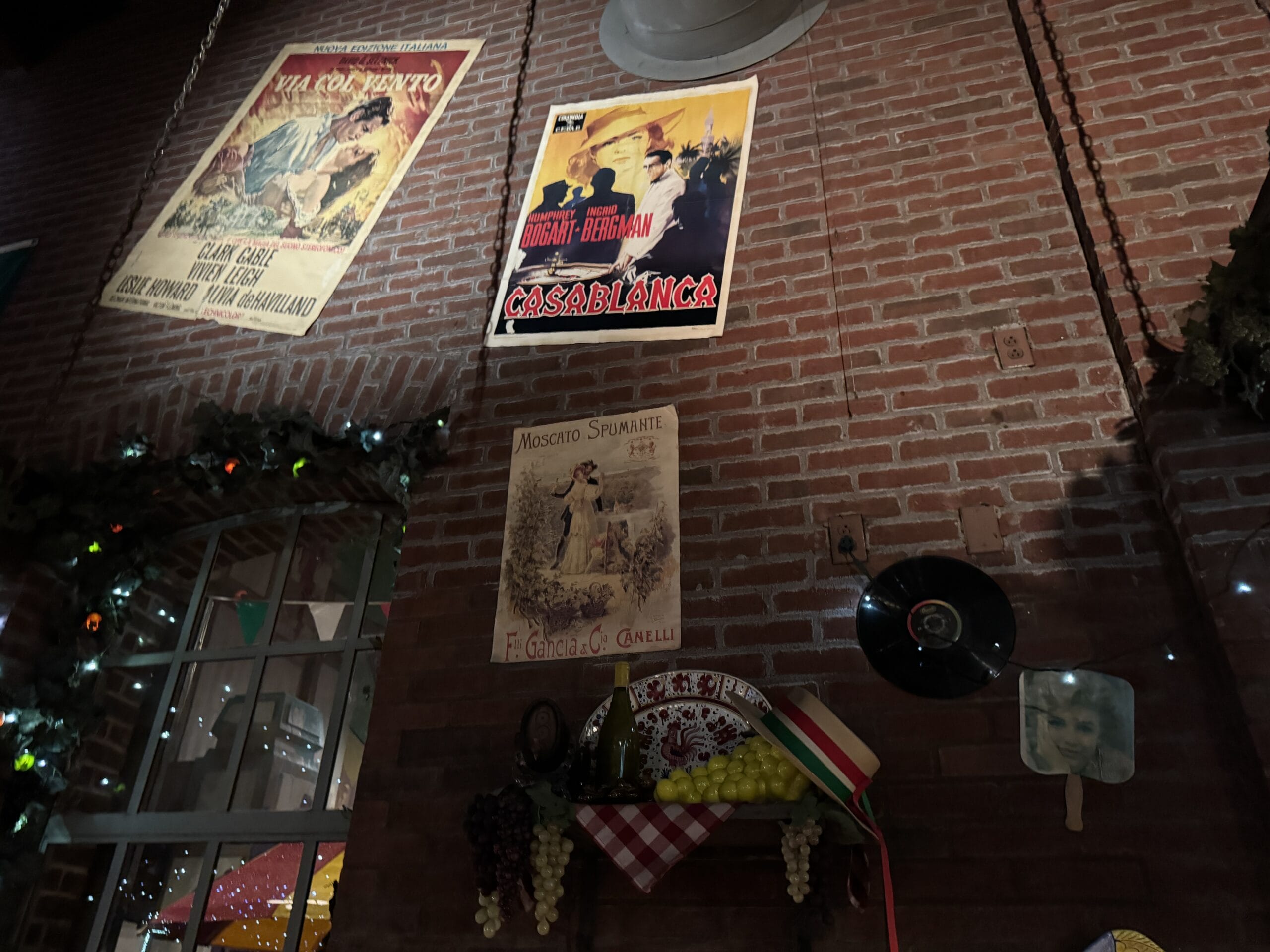 Brick wall at Mama Melrose’s Ristorante Italiano with vintage posters, record, grapes, string lights; window reflections on left.