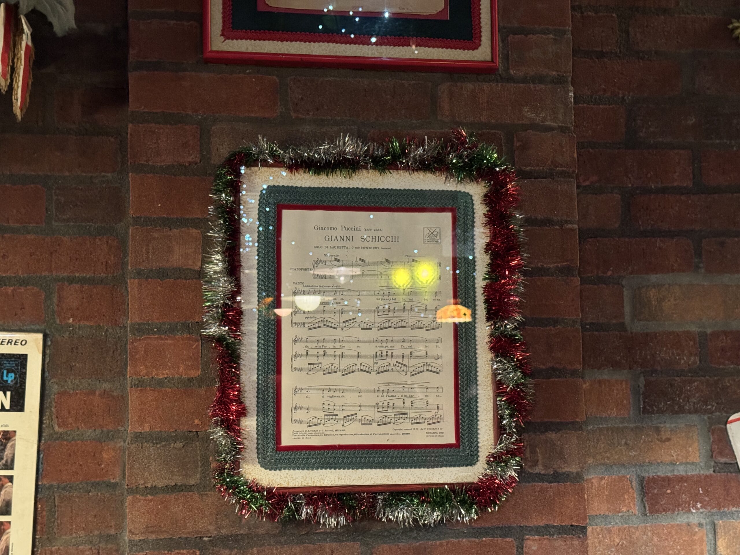 Framed sheet music at Mama Melrose’s Ristorante Italiano, adorned with festive tinsel, on a brick wall—perfect for photos.
