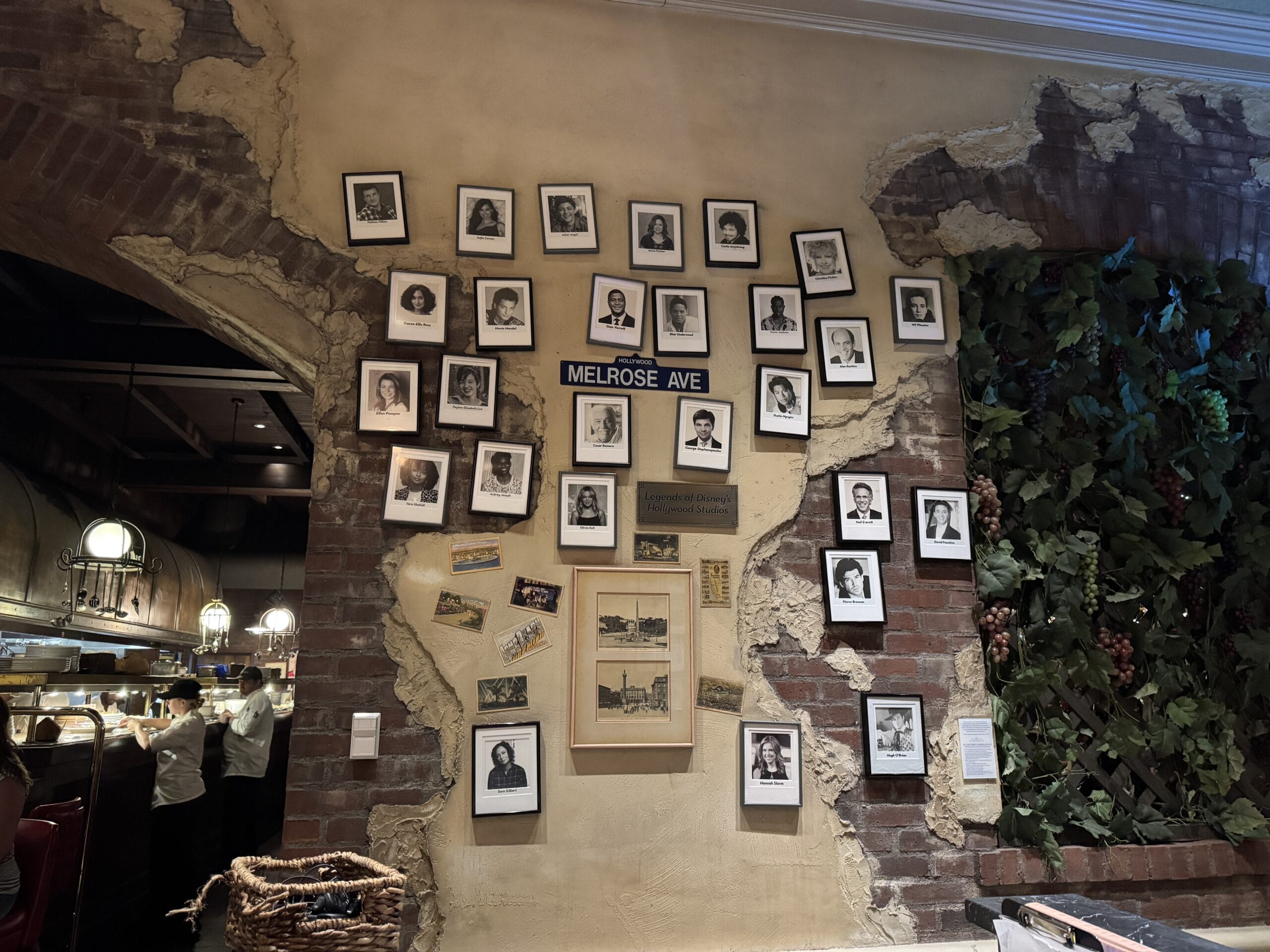 A rustic wall at Mama Melrose’s Ristorante Italiano with framed photos, a "Melrose Ave" sign, postcards, and kitchen staff.