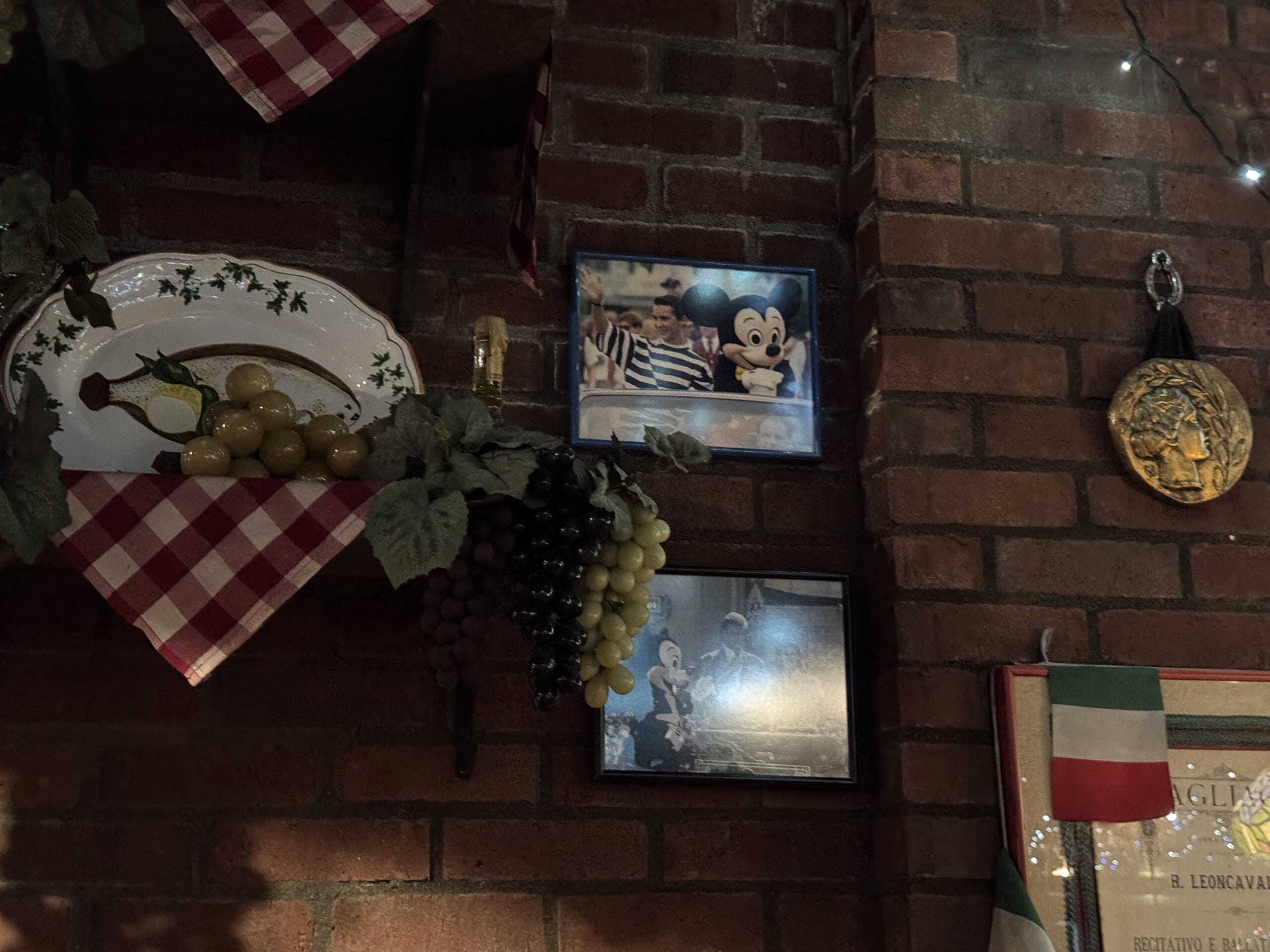 A brick wall at Mama Melrose’s Ristorante Italiano with framed photos, grapes, a medal, and Italian decor.