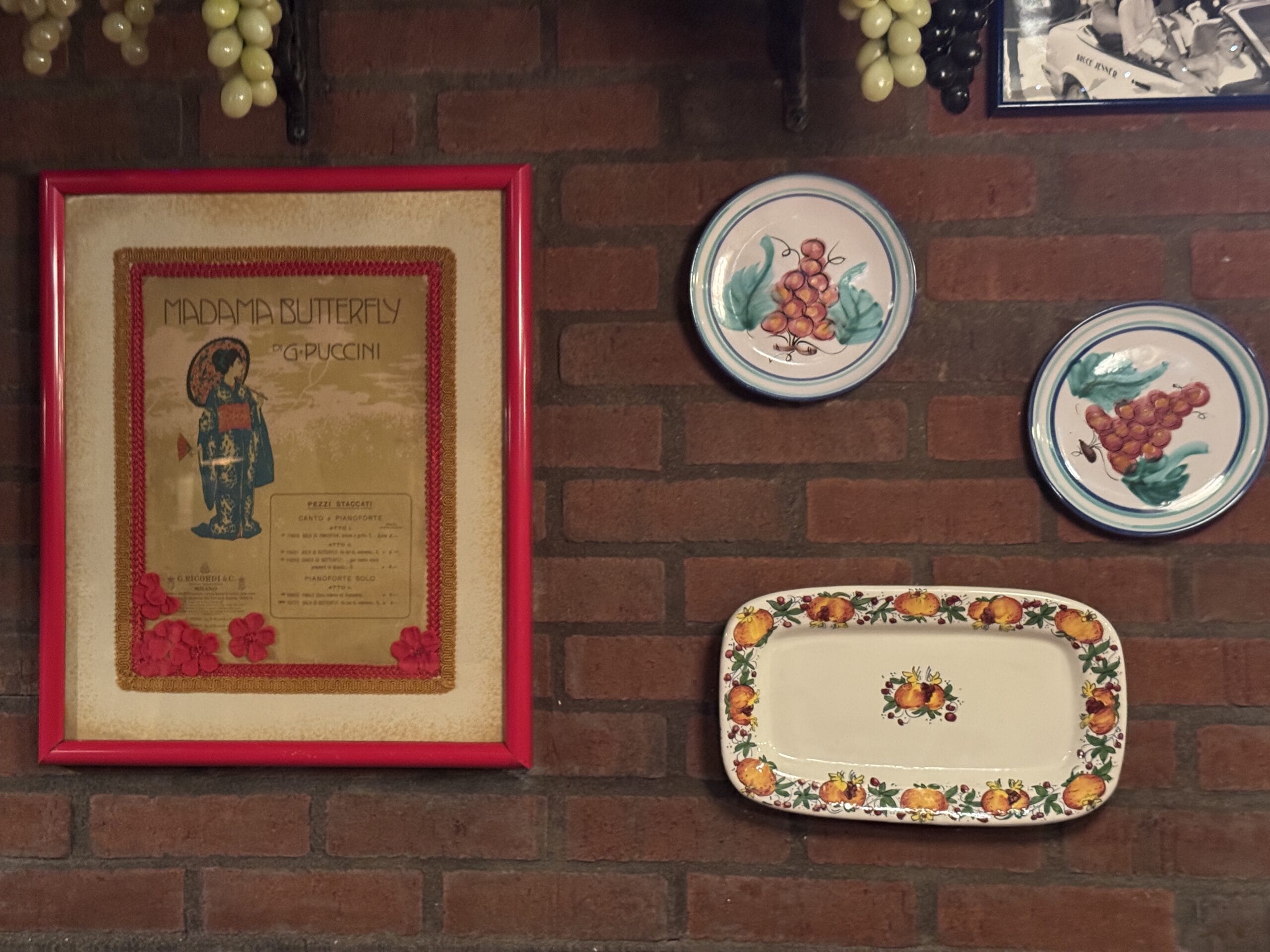 Framed "Madama Butterfly" poster and plates with grape designs on a brick wall at Mama Melrose’s Ristorante Italiano.