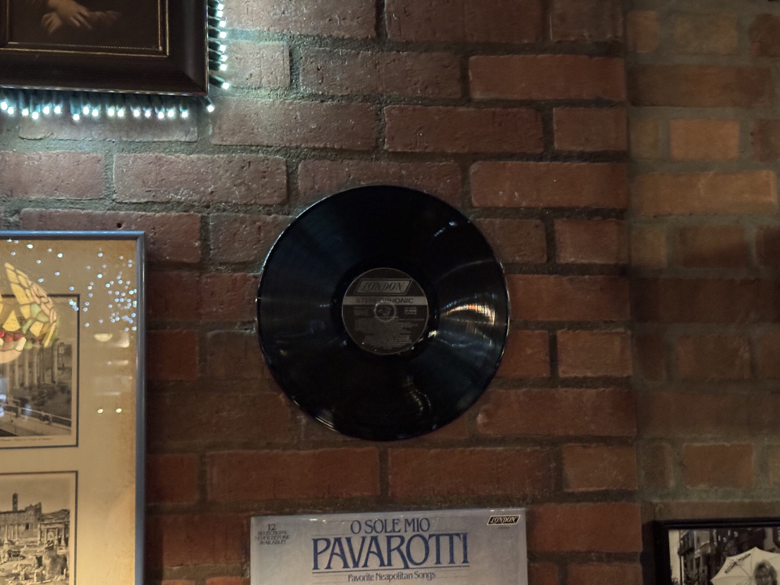 A black vinyl record is mounted on a brick wall at Mama Melrose’s Ristorante Italiano, surrounded by photos and posters.