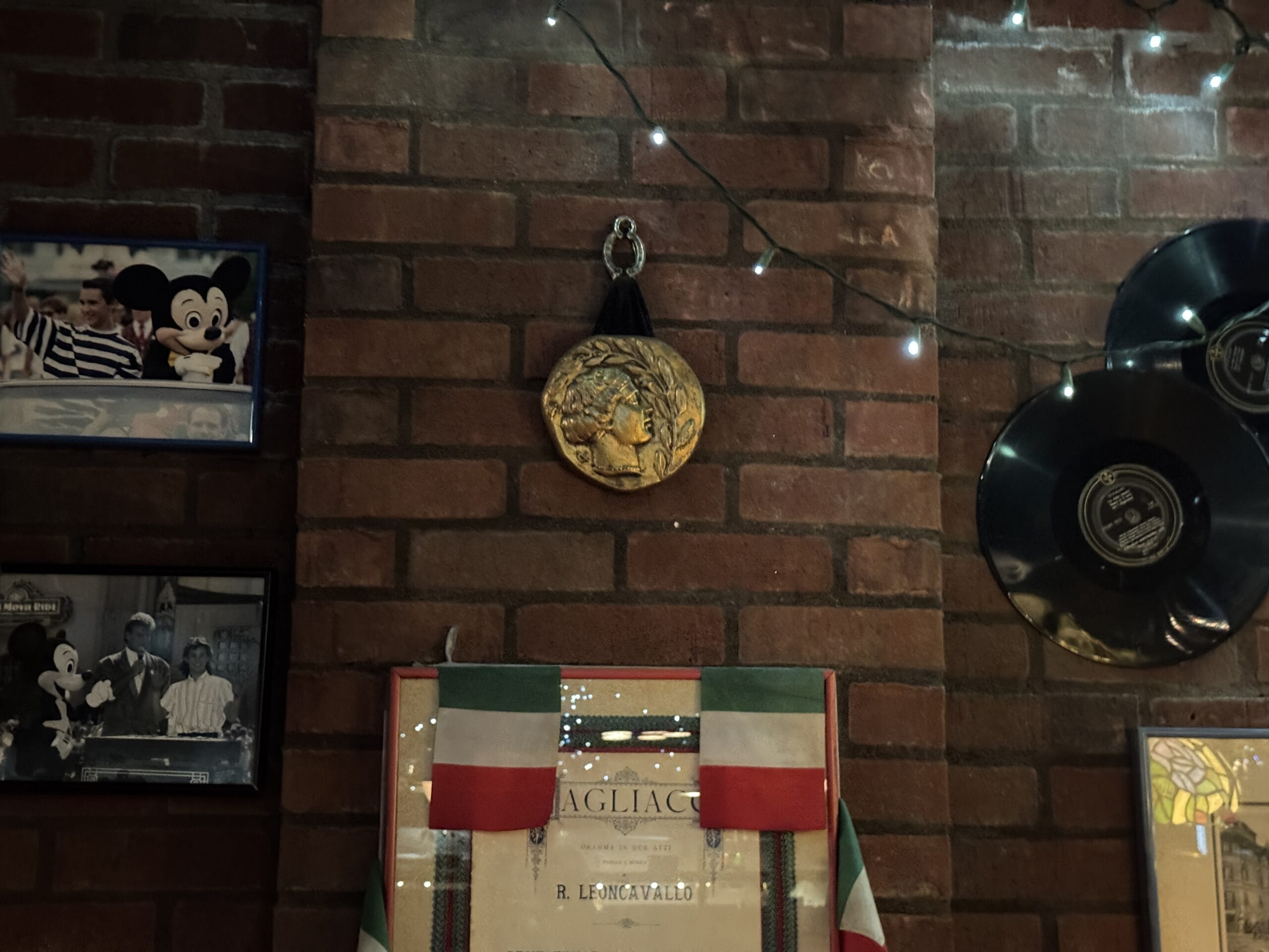 Gold medallion on brick wall at Mama Melrose’s Ristorante Italiano, with string lights, records, photos, and an Italian flag banner.