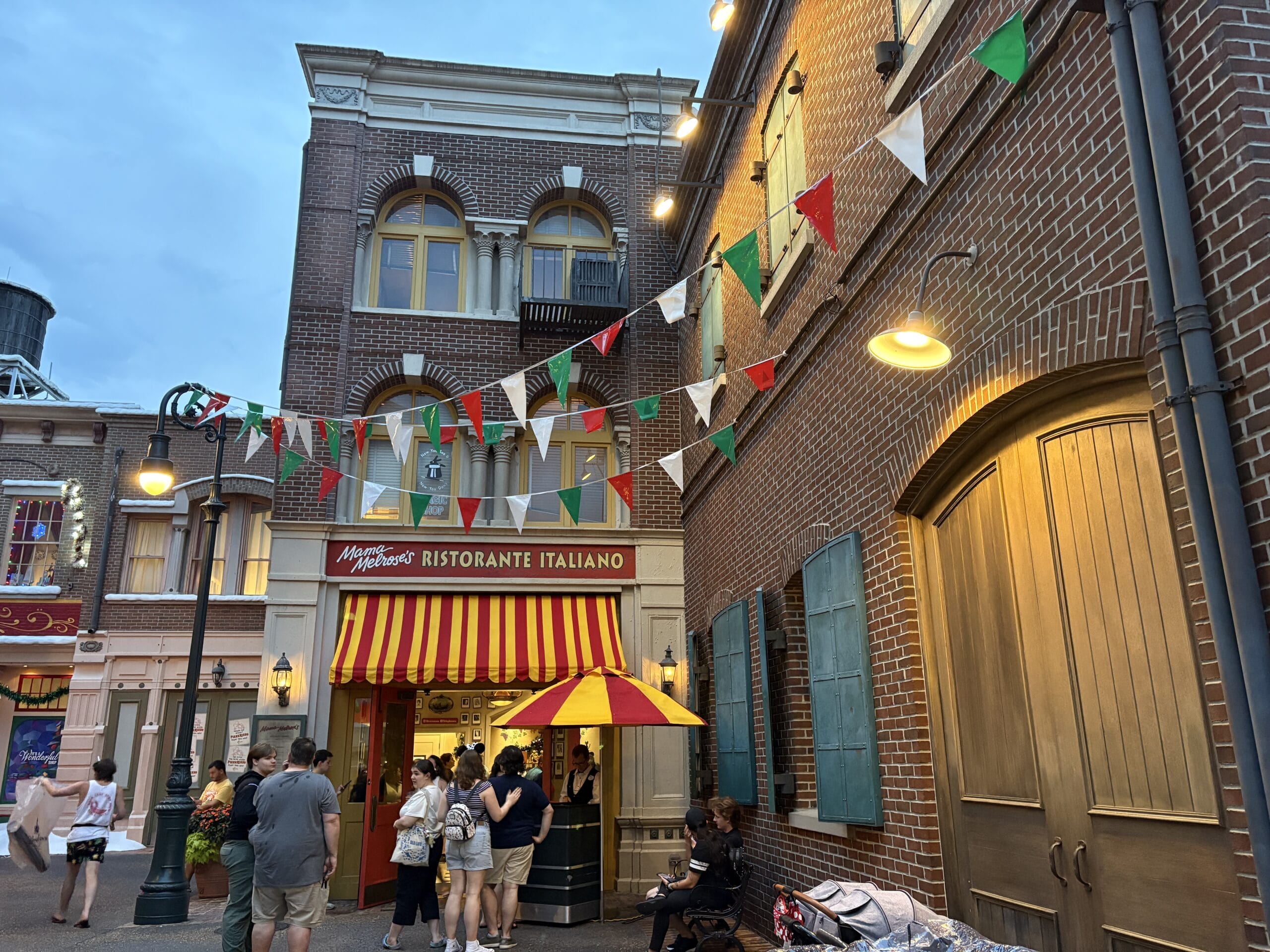 People pose for photos outside Mama Melrose’s Ristorante Italiano, beneath festive red, white, and green pennant banners.