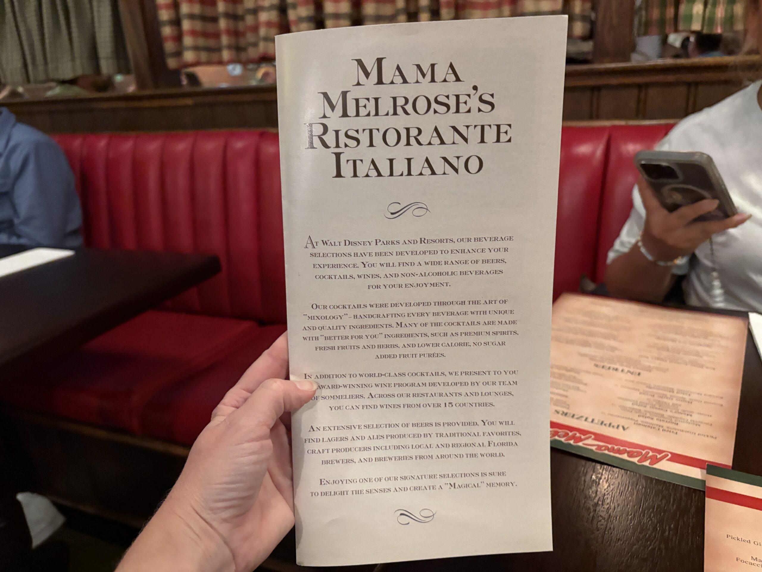 A hand holds a Mama Melrose’s Ristorante Italiano menu inside Disney’s Hollywood Studios; red booths and diners in the photo.