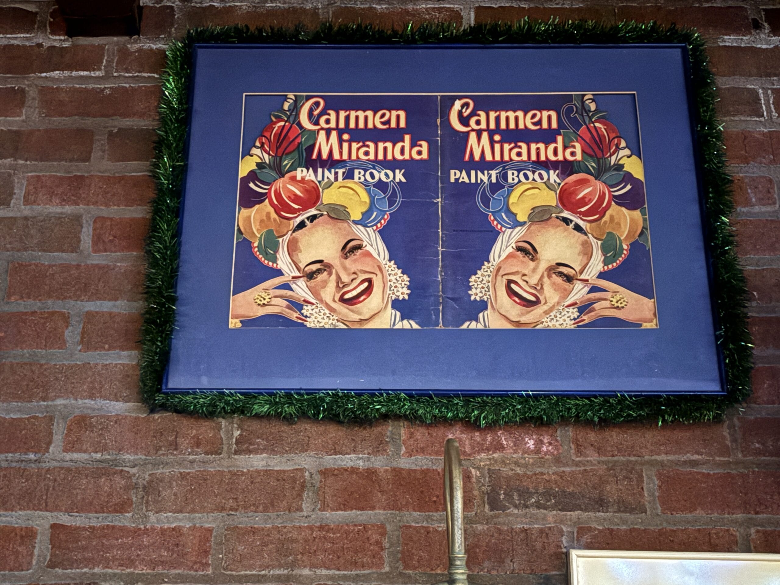 Framed vintage Carmen Miranda paint book cover with her smiling face and fruit hat, seen at Mama Melrose’s Ristorante Italiano.