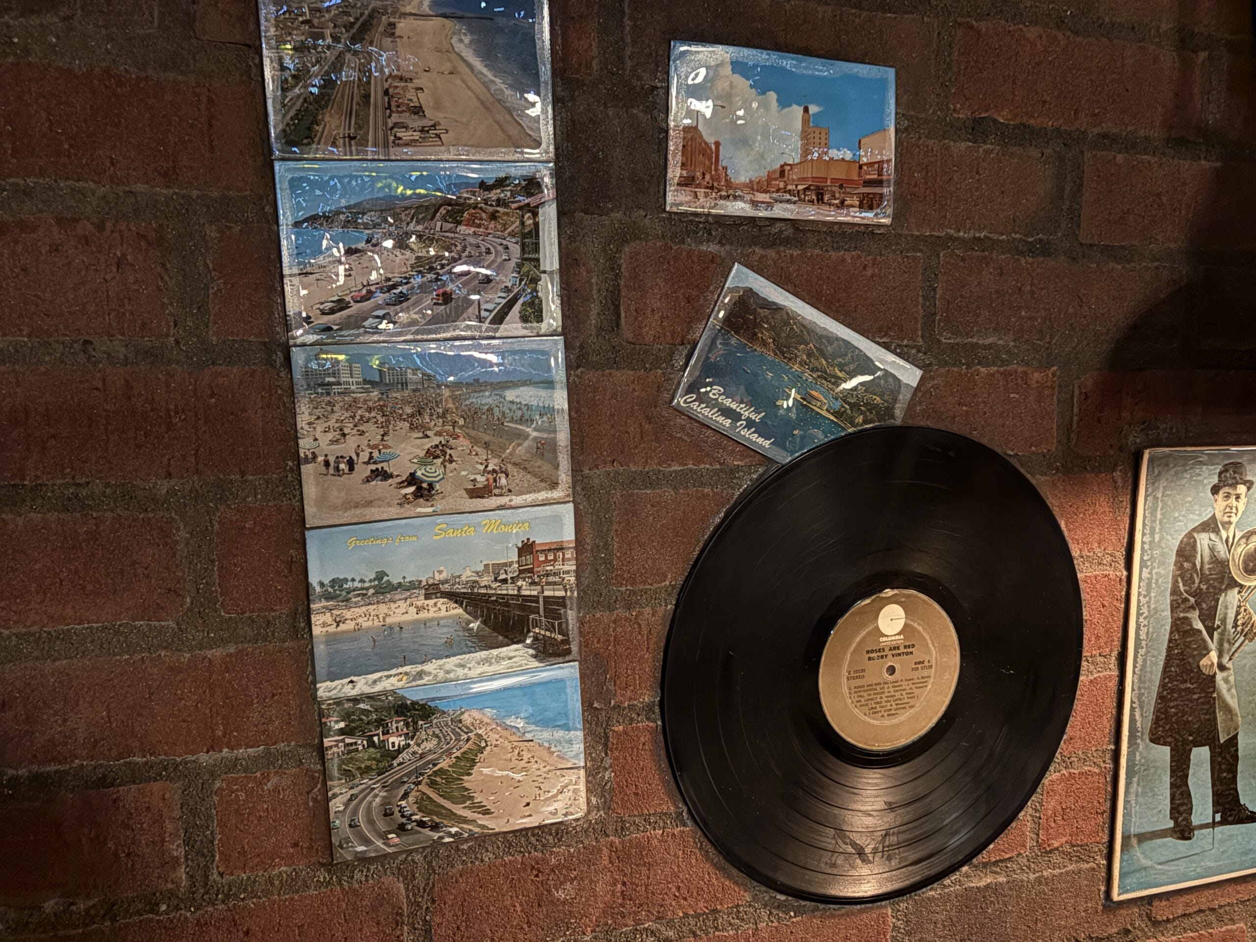 Vintage postcards, photos, and a vinyl record are displayed on a brick wall under warm light.