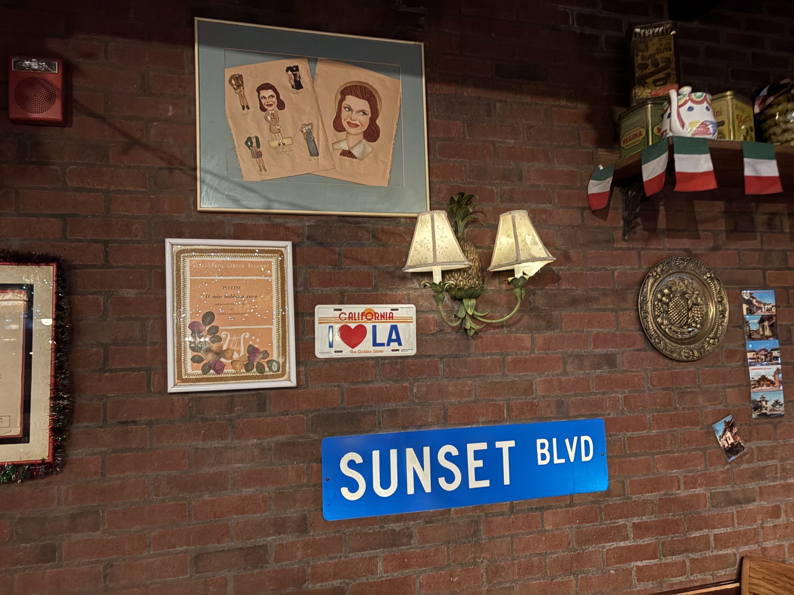Decorative brick wall with art, "SUNSET BLVD" sign, photos, and twin wall lamps at Mama Melrose’s Ristorante Italiano.