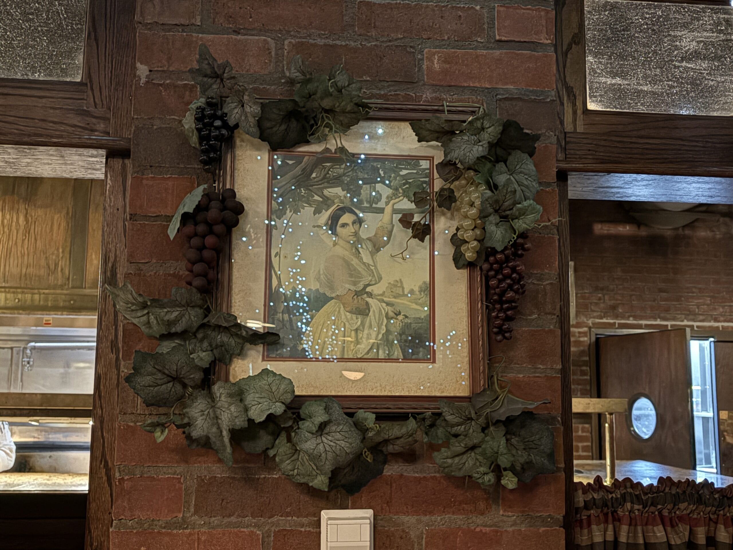 Framed painting of a woman with grapes, photos from Mama Melrose’s Ristorante Italiano on a brick wall.