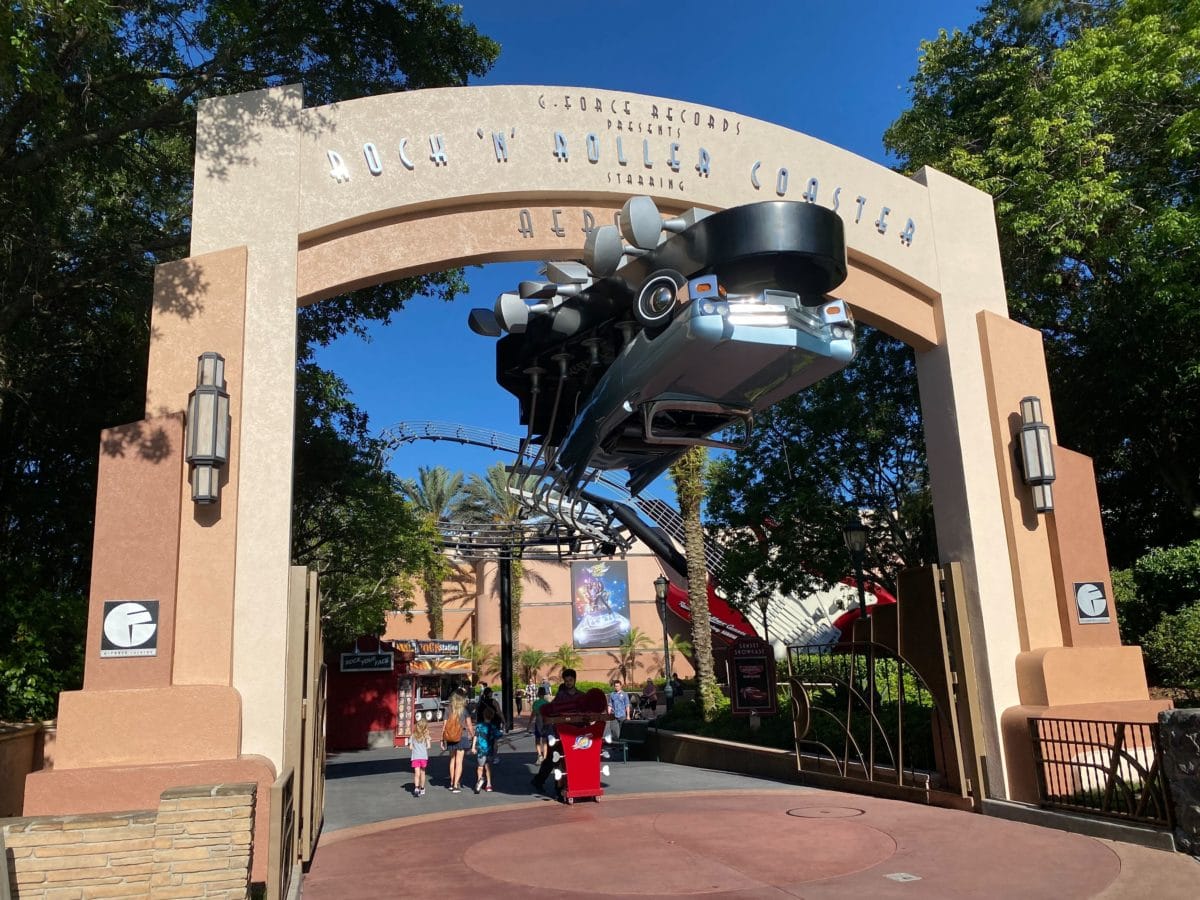 Large archway entrance to Rock n Roller Coaster, featuring a car sculpture and guests walking beneath, surrounded by trees.