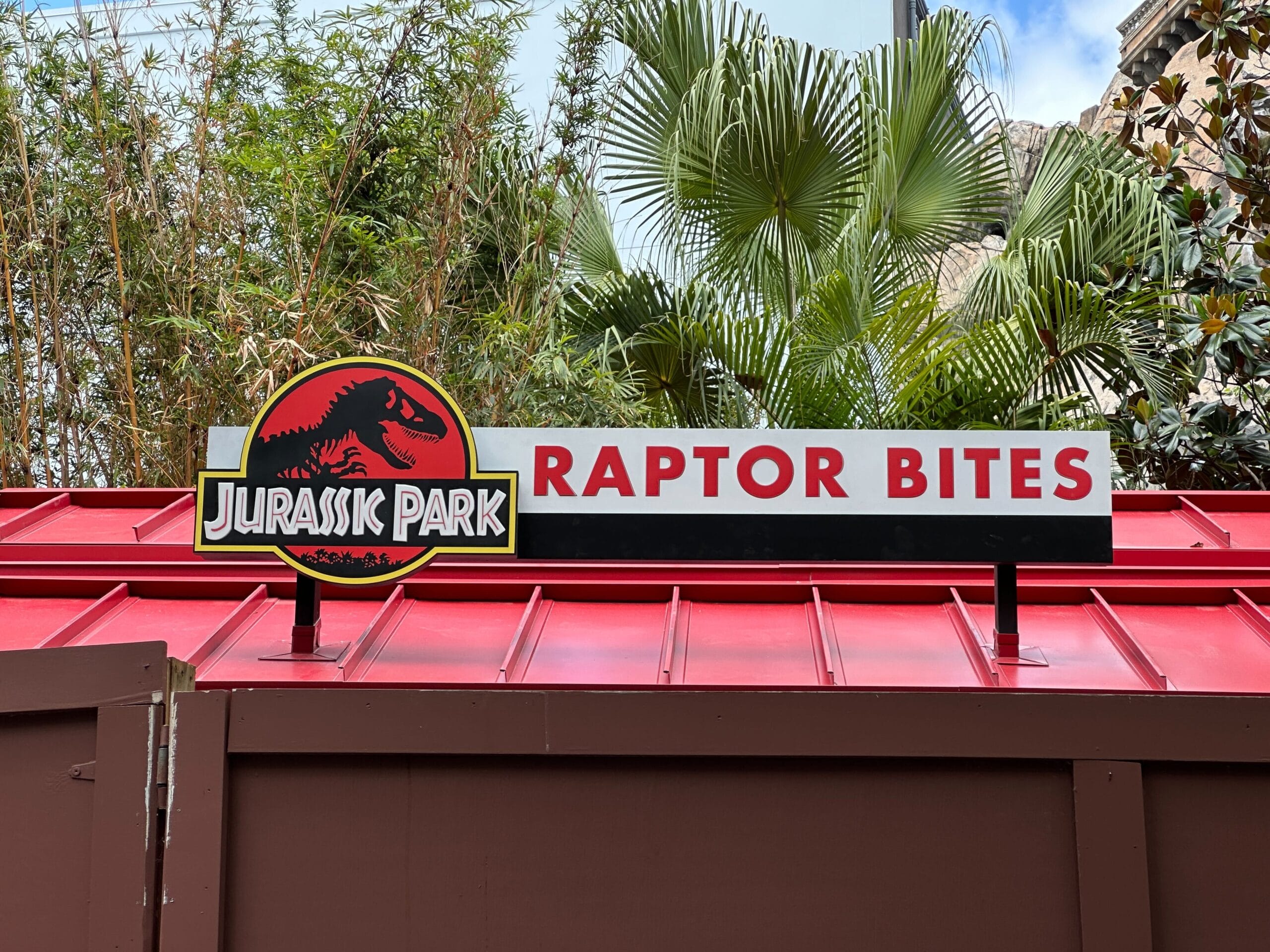 A "Jurassic Park Raptor Bites" sign on a red roof kiosk at Universal Islands of Adventure, framed by lush palm trees.