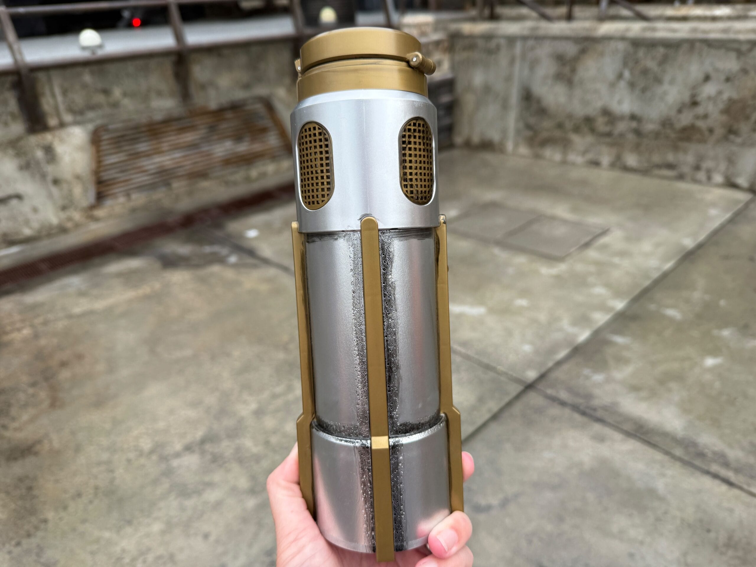 A hand holds a silver and gold cylindrical object with vents, against a concrete outdoor background.