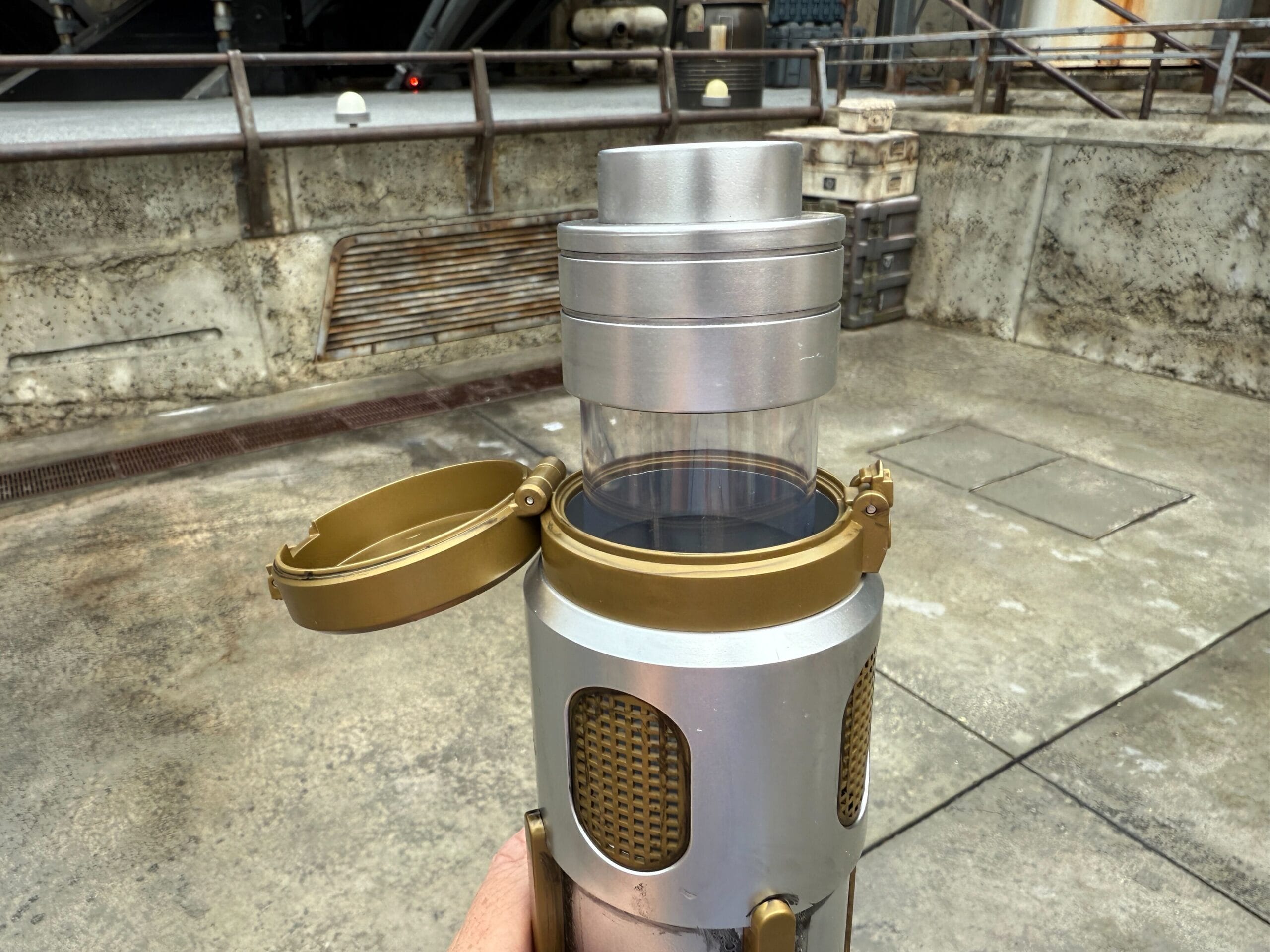 A hand holds a Coaxium Sipper with a gold lid open, evoking Star Wars Day against an industrial concrete background.