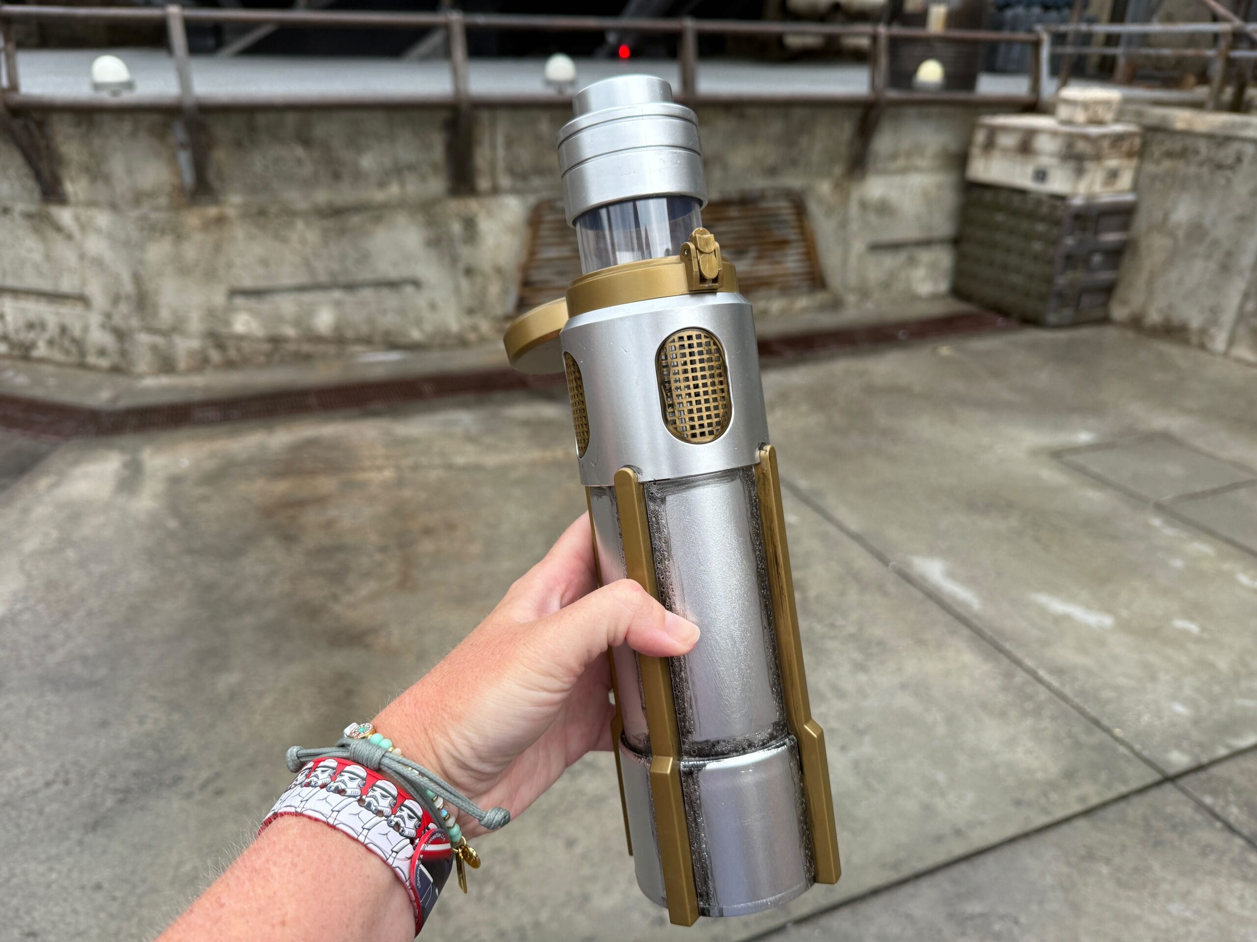 A hand holds a silver and gold Coaxium Sipper, resembling a custom lightsaber, outdoors at Walt Disney World.
