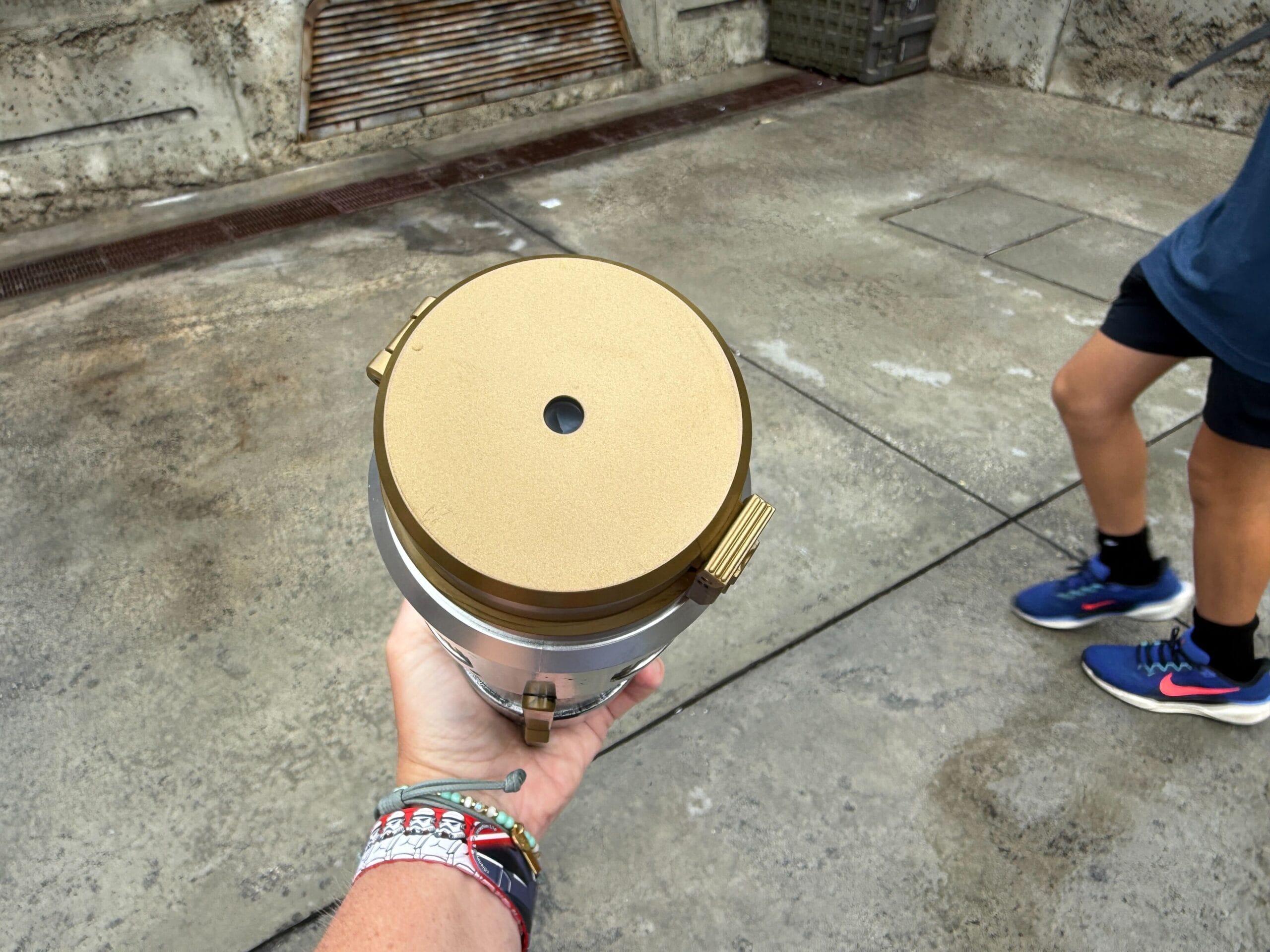 A person holds a golden cylindrical container outdoors, with another person's legs visible walking by on the right.