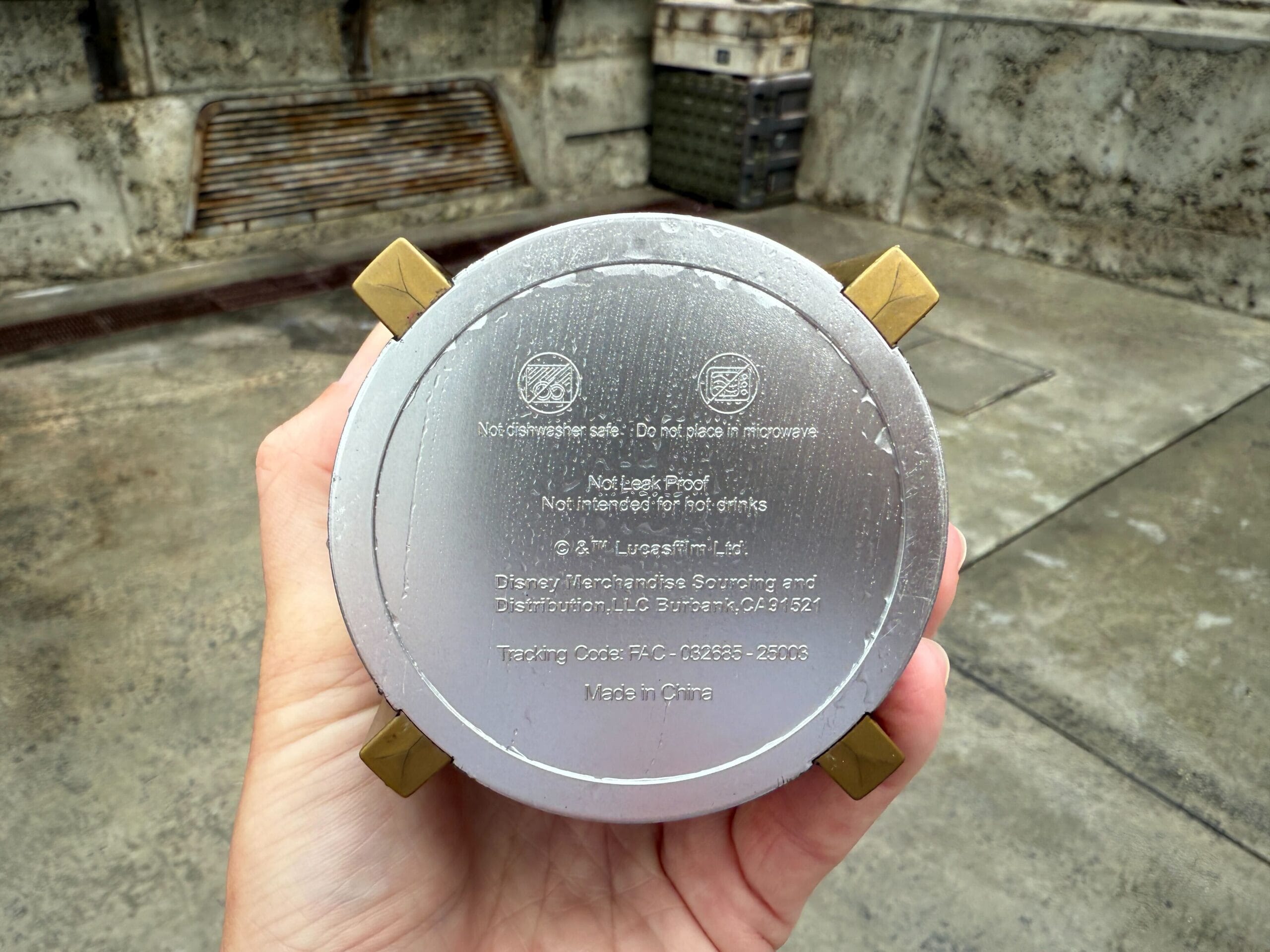 A hand holds a silver cup upside down, showing product warnings and manufacturing details on the base.