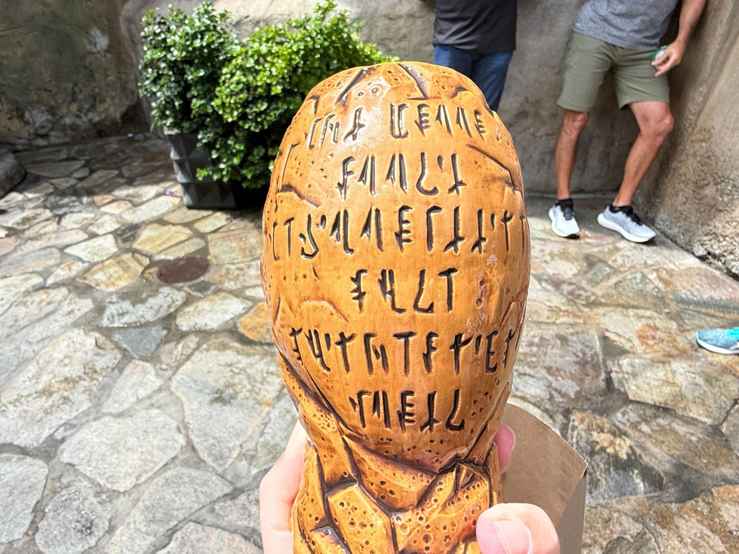 A hand holds a carved object with runic writing; people stand in the background on a stone-paved surface.