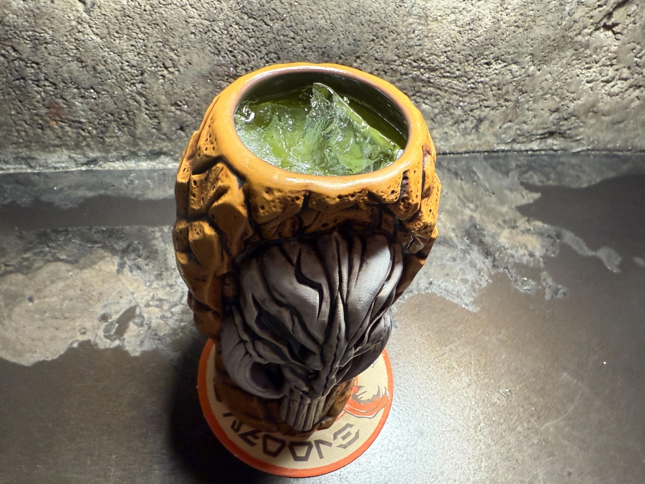 A tiki-style skull mug filled with a green iced drink sits on a coaster against a textured gray background.