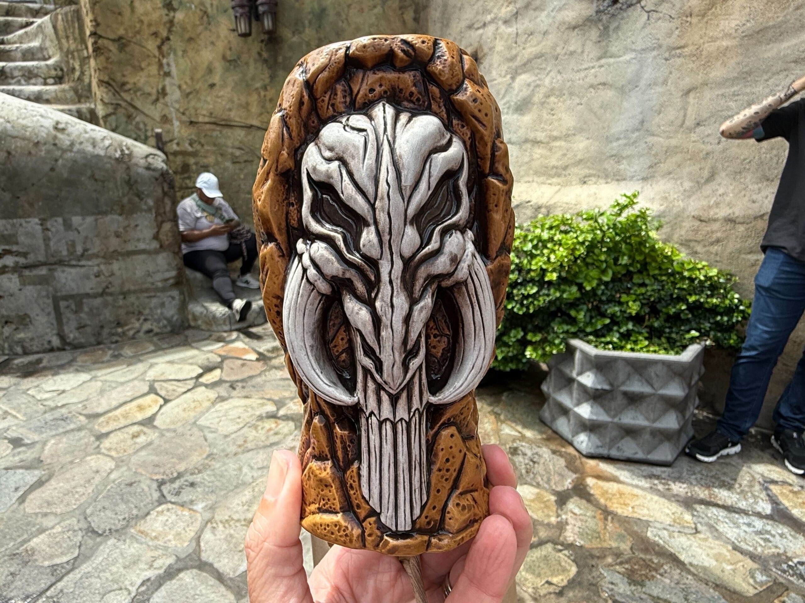 A hand holds a carved cup featuring a detailed skull with large tusks; stone paths and plants are in the background.