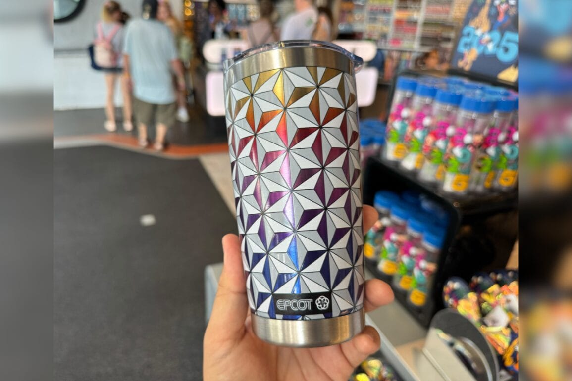 A hand holds a Metallic Spaceship Earth Tumbler in a colorful EPCOT store with people in the background.