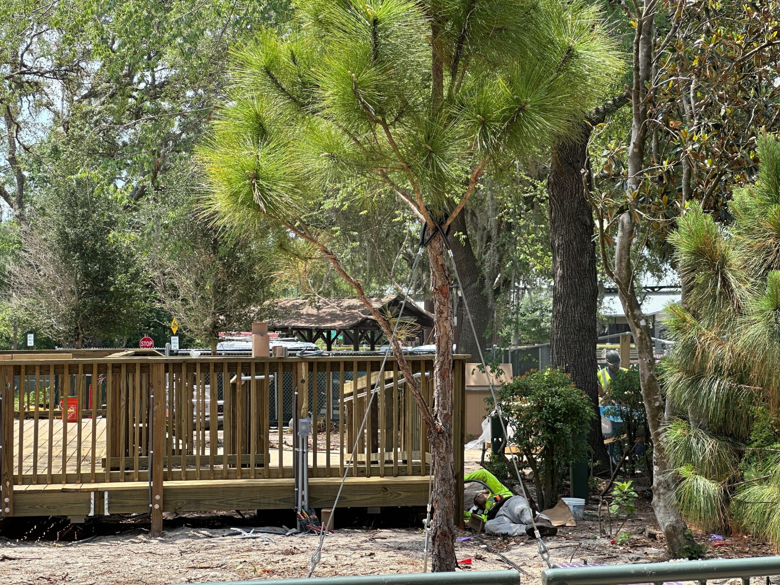 A worker in a neon vest kneels by a wooden deck, assisting with cabin construction amid trees and greenery on a sunny day.