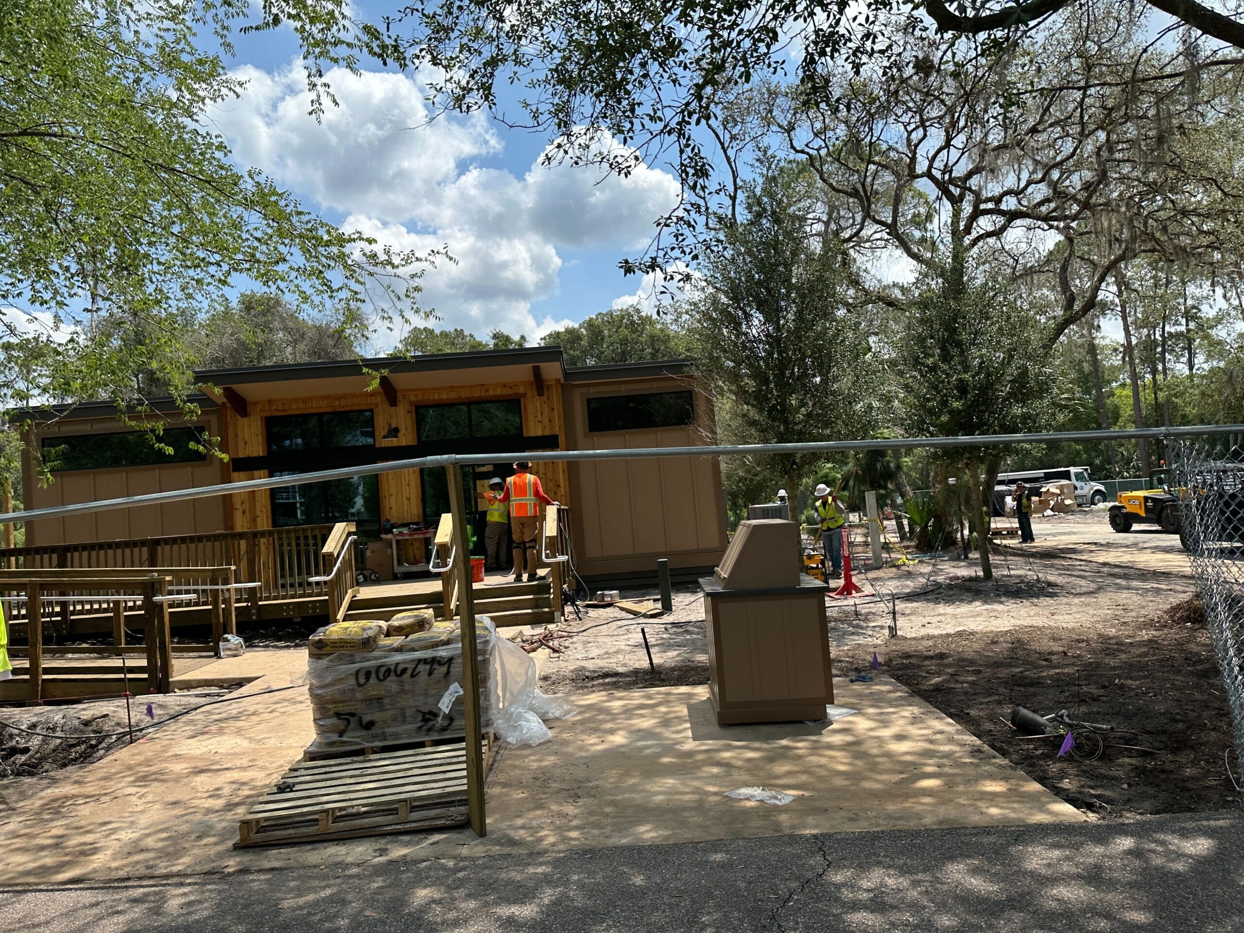 Cabin builders in safety vests work outside a building surrounded by trees, with equipment and materials nearby.