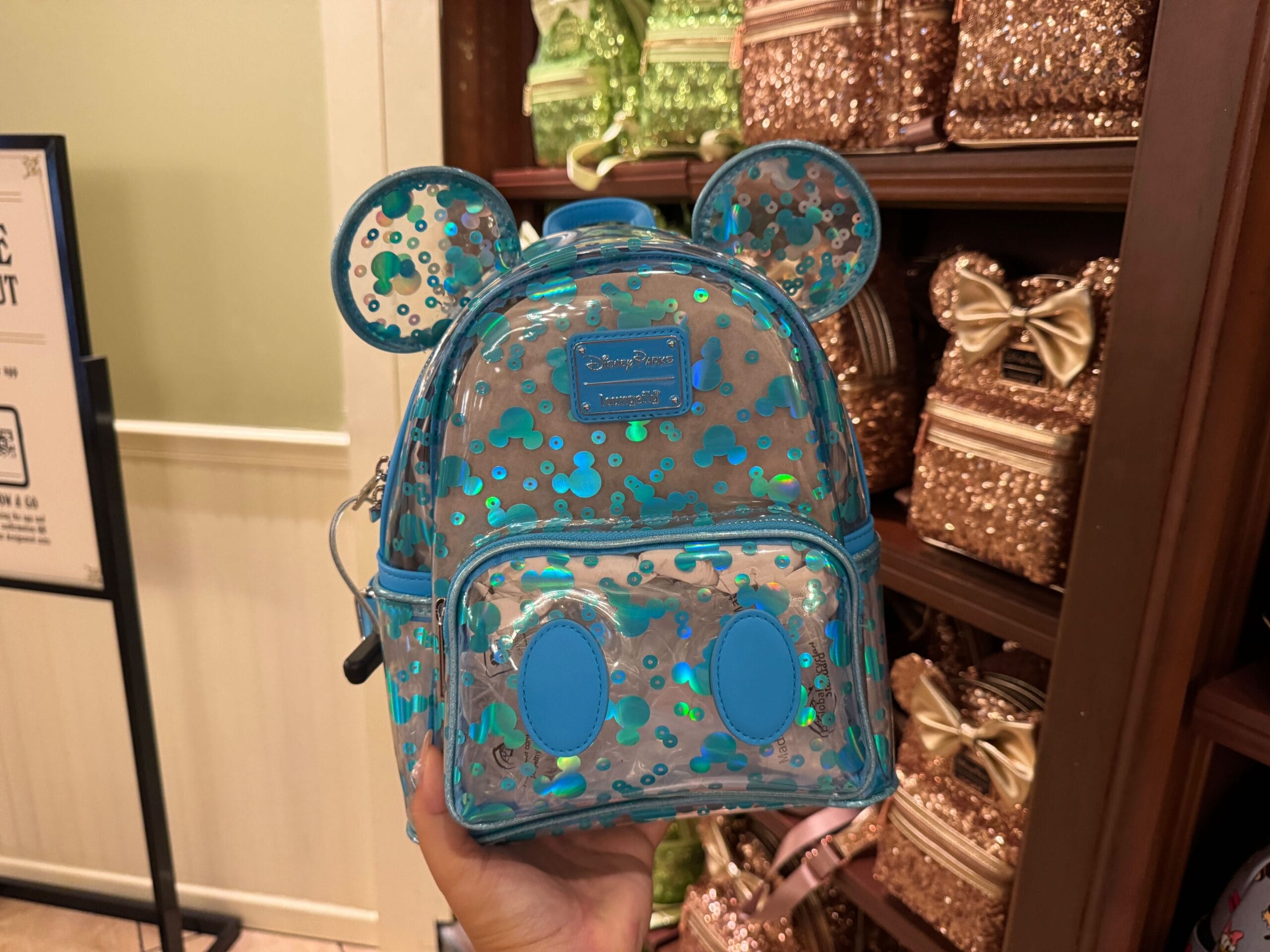 A hand holds a clear loungefly mini backpack with mouse ears and blue glitter spots, displayed in a store.