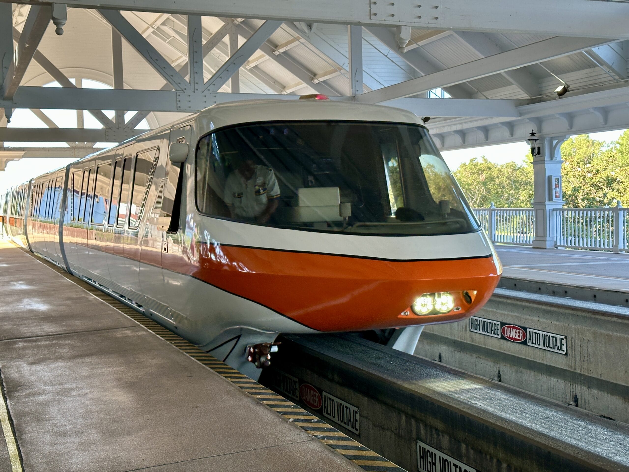 A sleek, orange and white monorail arrives at a covered outdoor station in Walt Disney World’s Magic Kingdom.