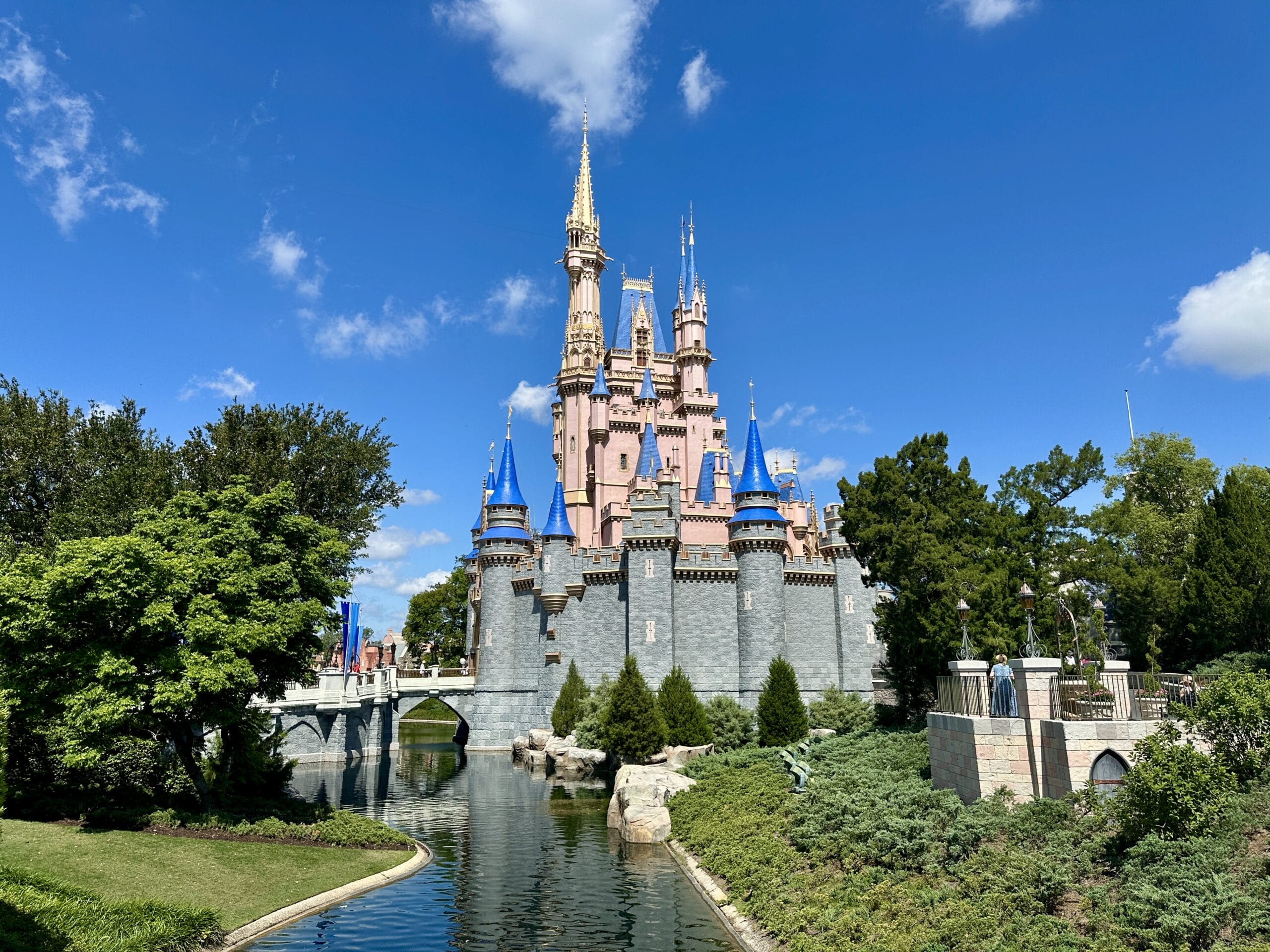 Cinderella Castle at Magic Kingdom, with the Liberty Belle Riverboat nearby, under a bright blue sky and clouds.