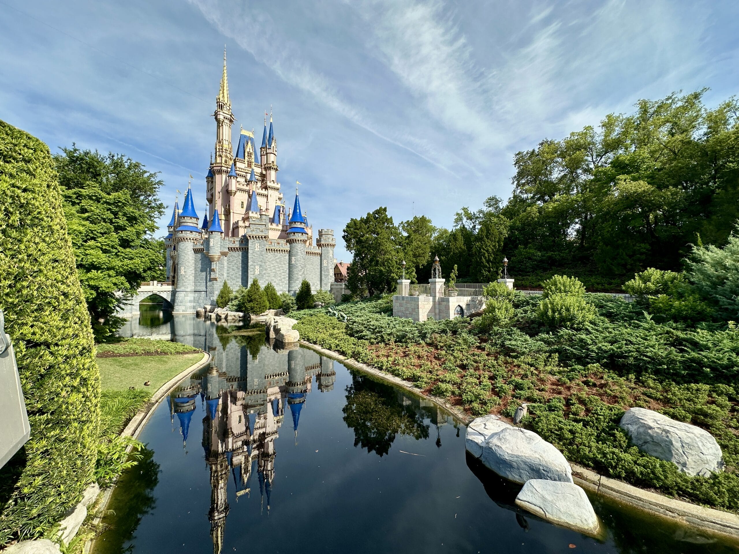 Cinderella Castle at Magic Kingdom with blue turrets reflected in a pond, surrounded by greenery under a blue sky.