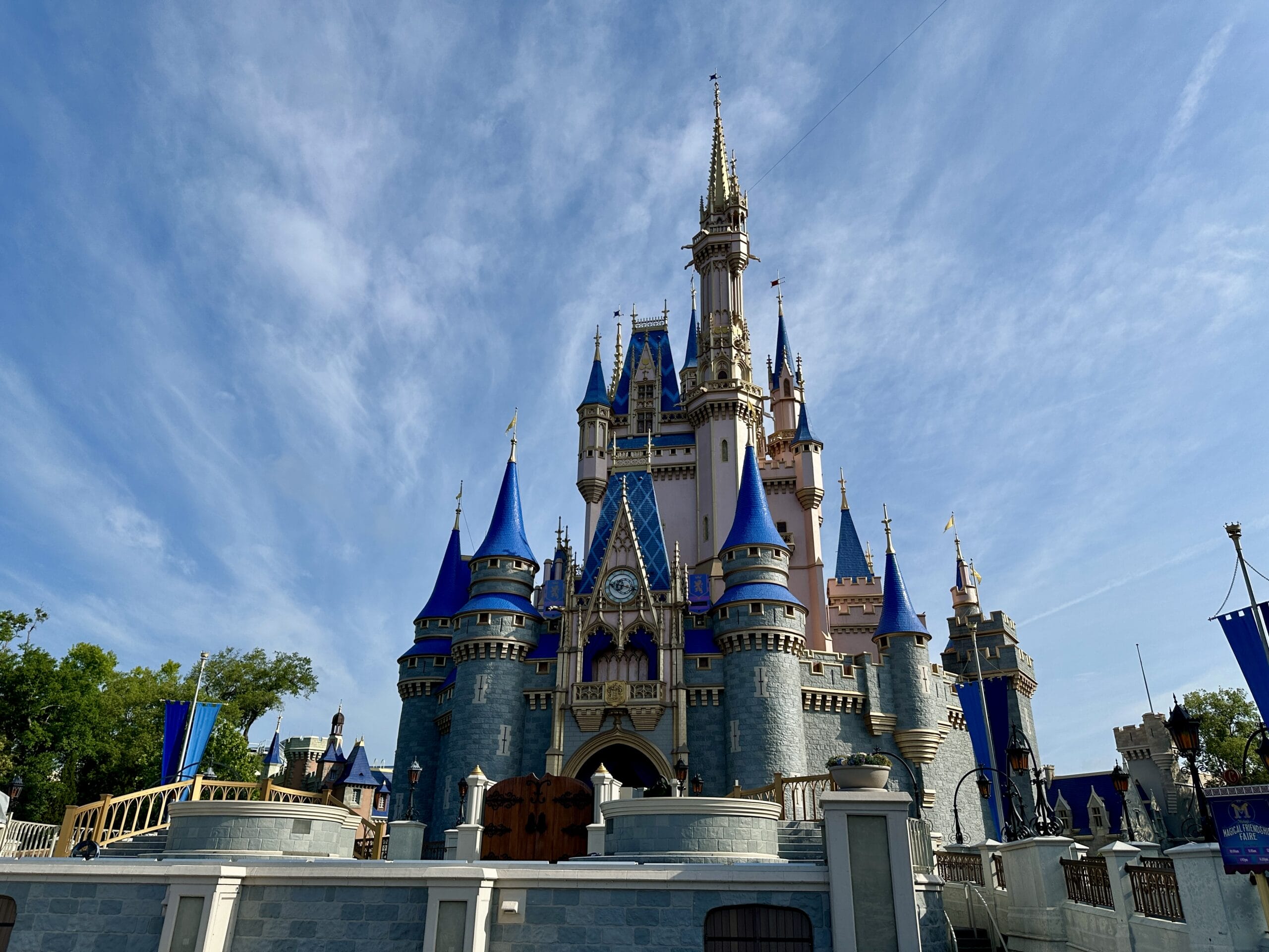 A fairytale castle with blue rooftops and tall spires shines under Disney World's bright sky with wispy clouds.