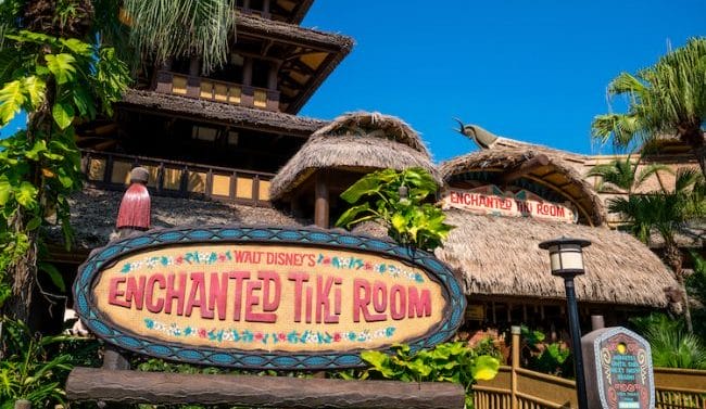 Entrance to Walt Disney’s Enchanted Tiki Room in Adventureland, with a colorful sign and tropical decor under a clear blue sky.