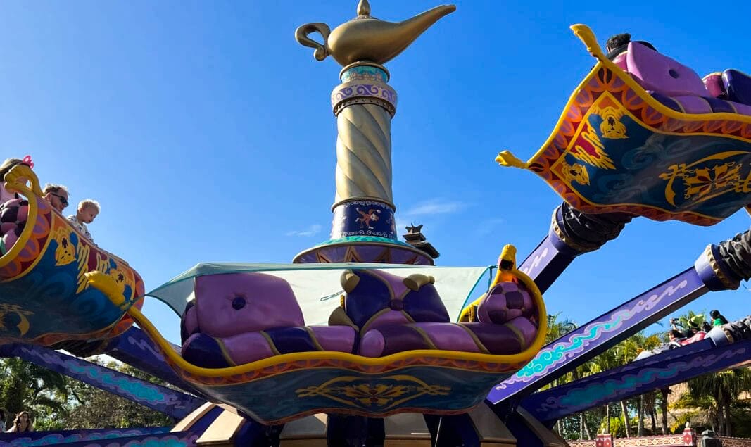 Children soar on flying purple magic carpets around a giant genie lamp in Adventureland under a clear blue sky.
