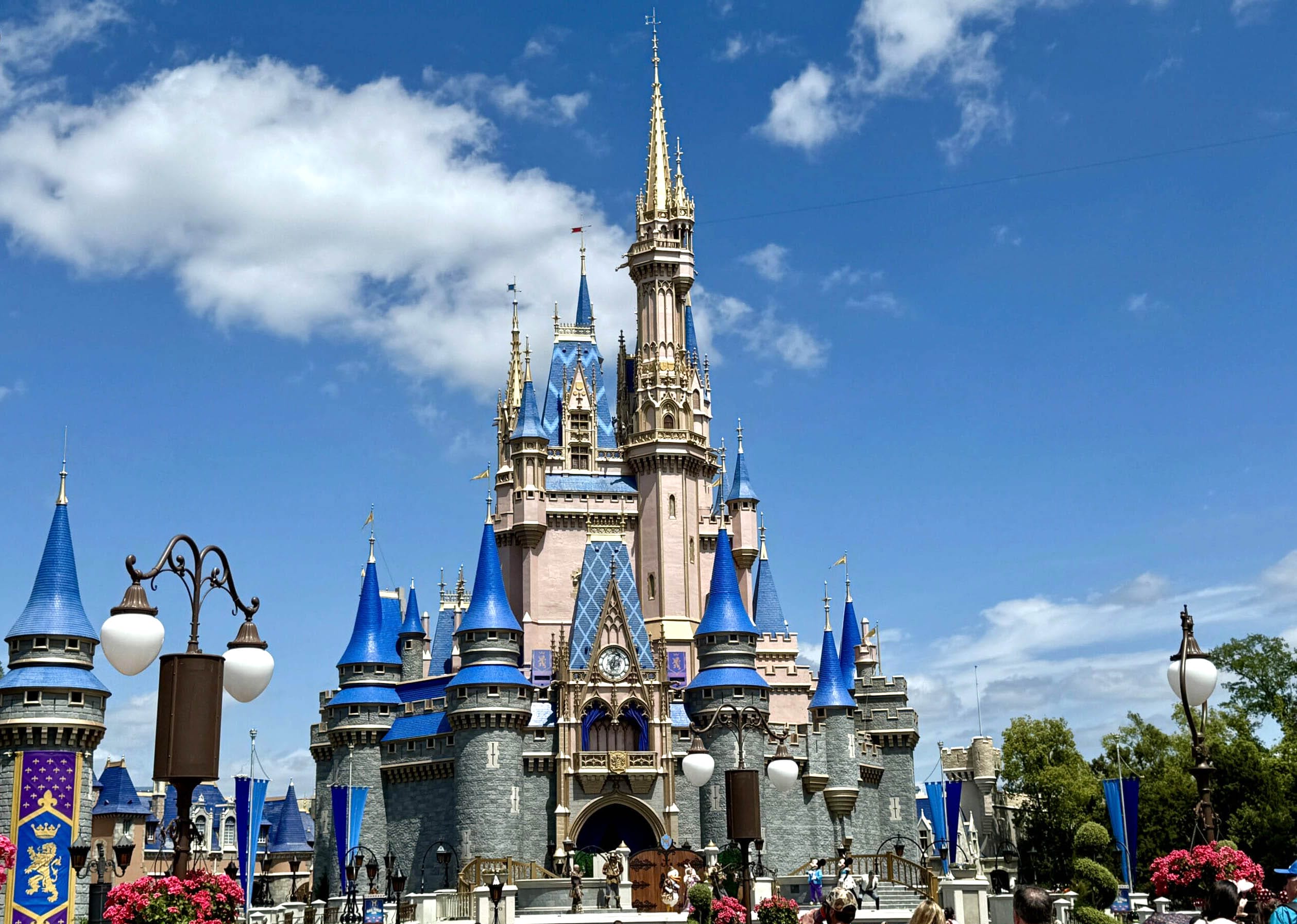 Cinderella Castle at Magic Kingdom, with blue spires and gold accents, under a bright blue sky with clouds.
