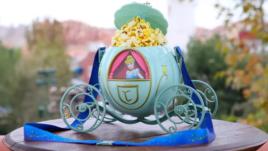 Blue Disney Popcorn Bucket shaped like Cinderella's carriage, filled with popcorn on a round wooden table outdoors.