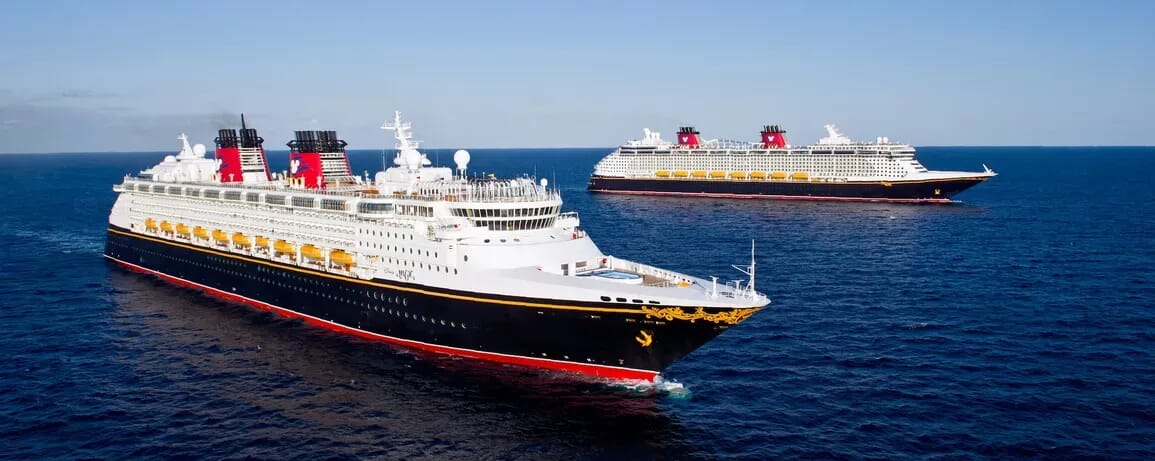 Two large Disney Cruise Line ships with red funnels sail on a calm blue ocean under a clear sky.