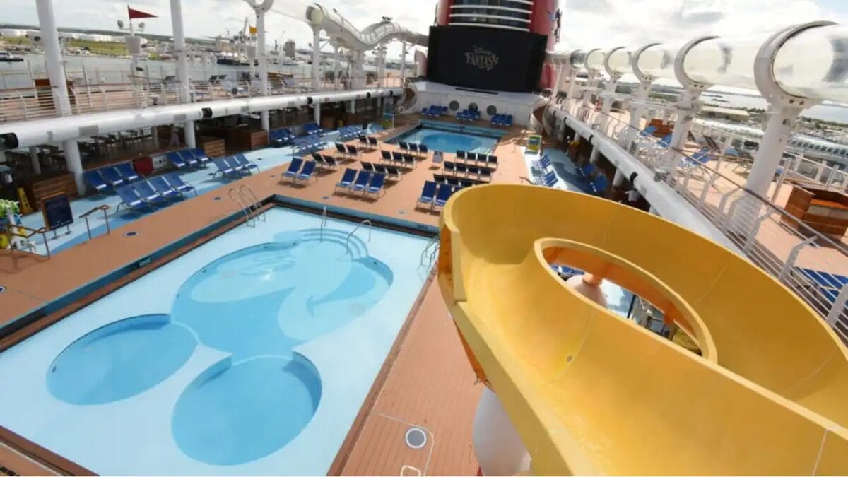 Disney Cruise ship pool deck with a yellow slide, blue lounge chairs, and a Mickey Mouse-shaped pool.