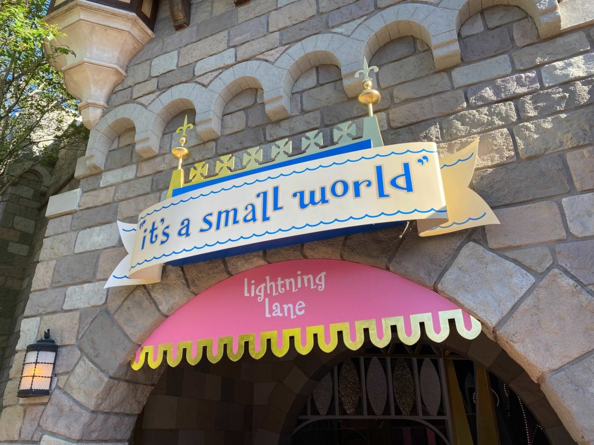 “it's a small world” entrance sign above a pink Disney Lightning Lane awning at a stone building in Disneyland.