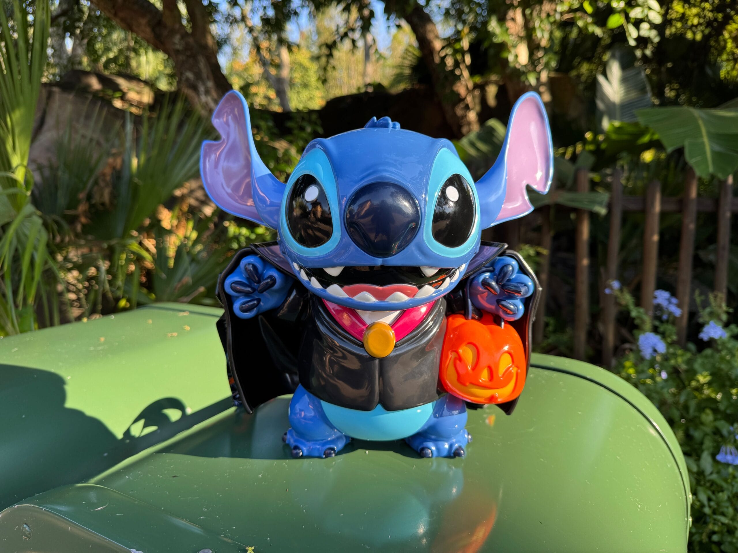 A Stitch figurine dressed as a vampire holds a Disney Popcorn Bucket, standing on a green surface outdoors.