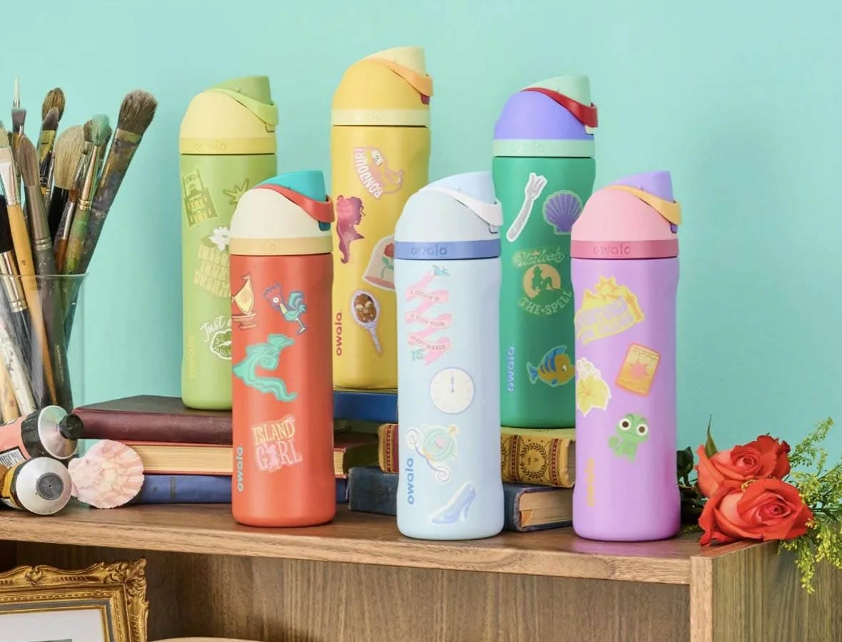 Five colorful Owala Collection water bottles with playful designs sit on a shelf beside books, paintbrushes, and red roses.