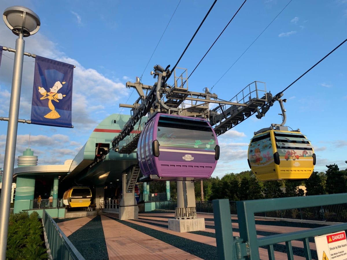 Purple and yellow Disney Skyliner gondolas arrive at an outdoor cable car station under a blue sky.