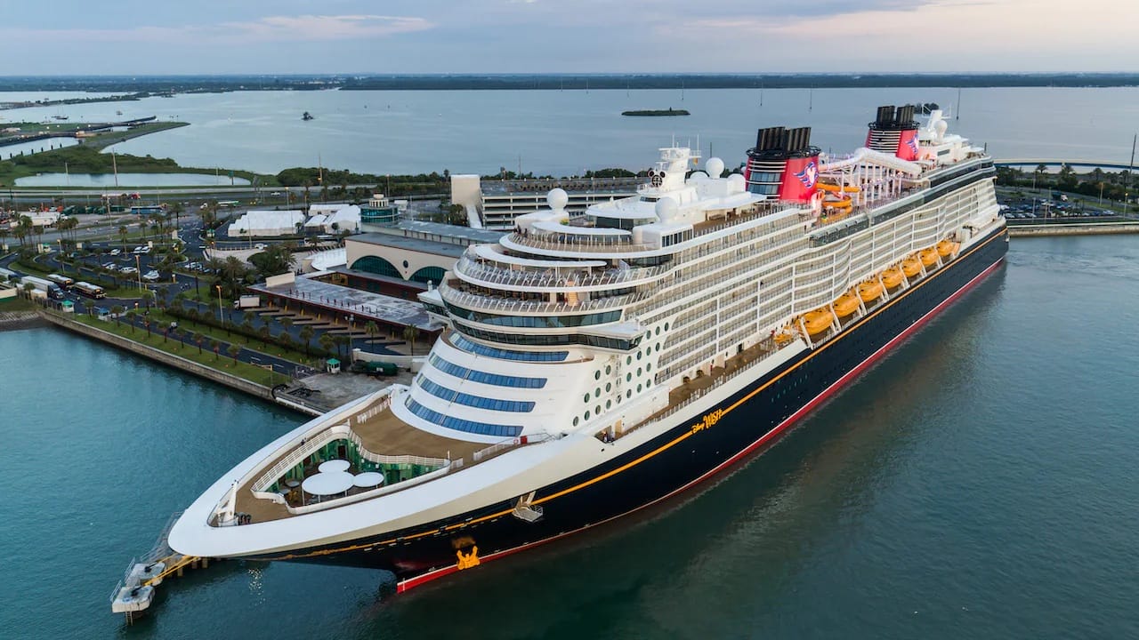 A Disney Cruise Line ship docked at a port with water and land in the background under a cloudy sky.
