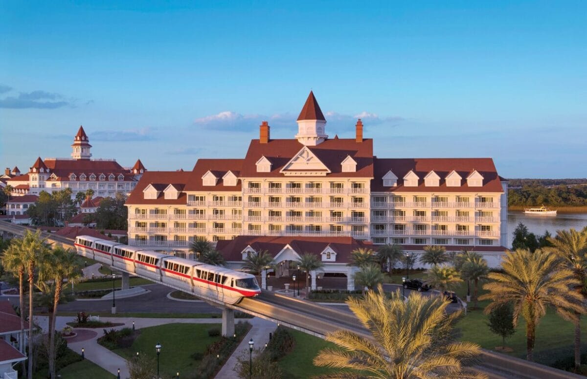 A white monorail glides past a Victorian-style Disney World Grand Floridia Resort with red roofs and palms.