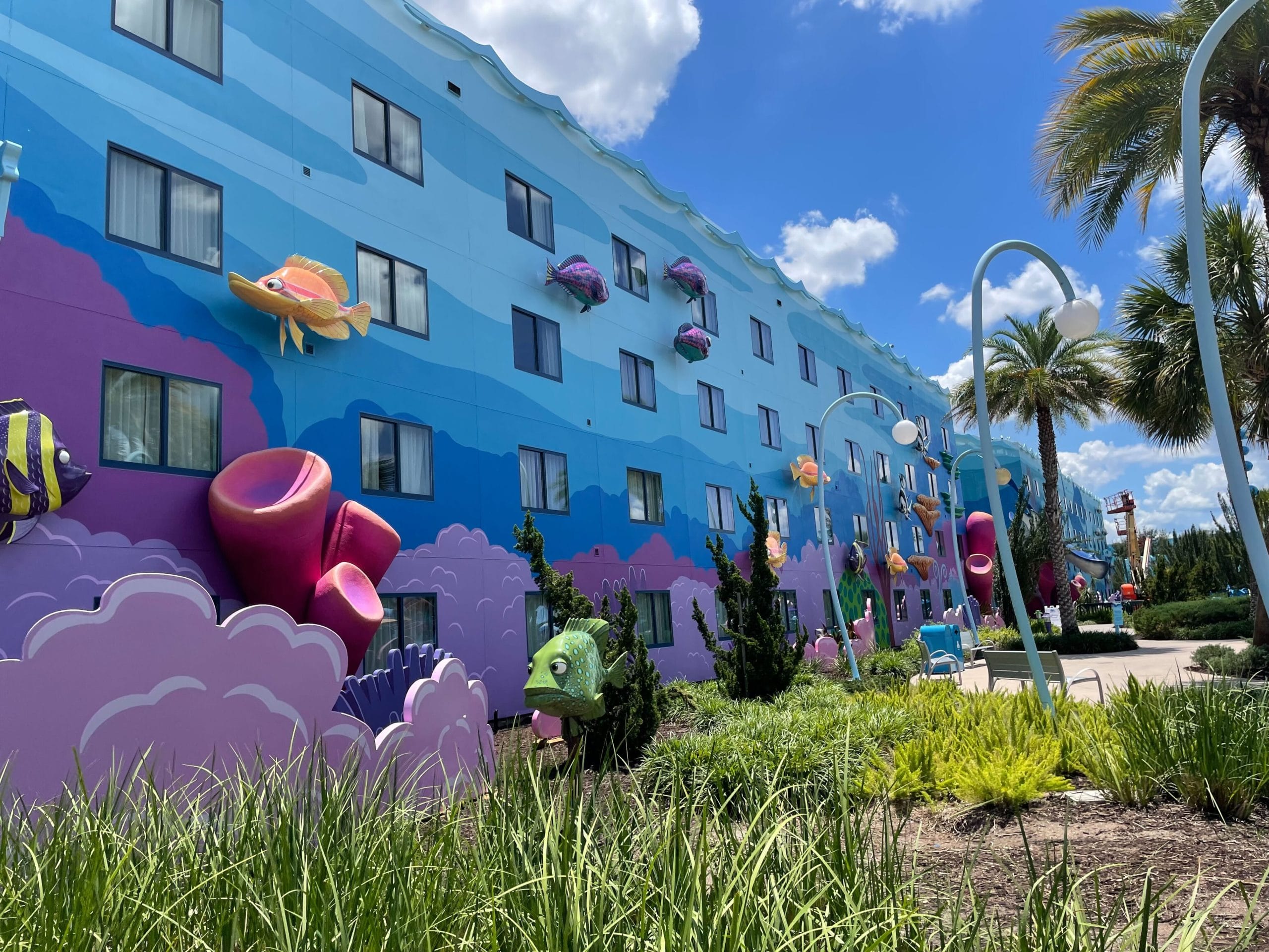 Colorful Disney World Art of Annimation hotel with whimsical sea-themed decor, fish, coral, palm trees, and greenery under a blue sky.