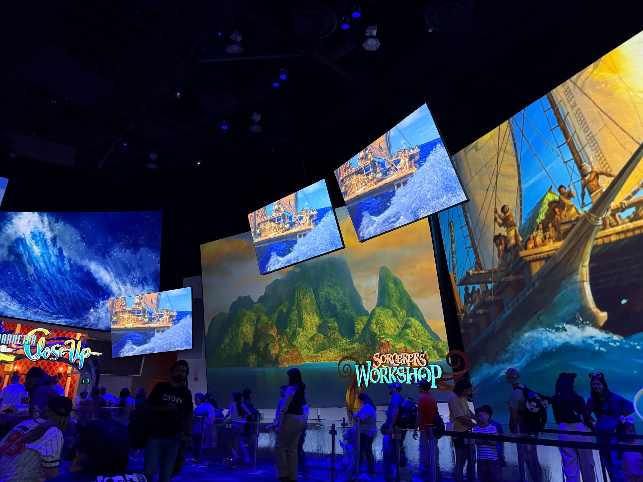 Colorful indoor exhibit with large screens showing ocean and island scenes, and a sign reading “Sorcerer’s Workshop.”