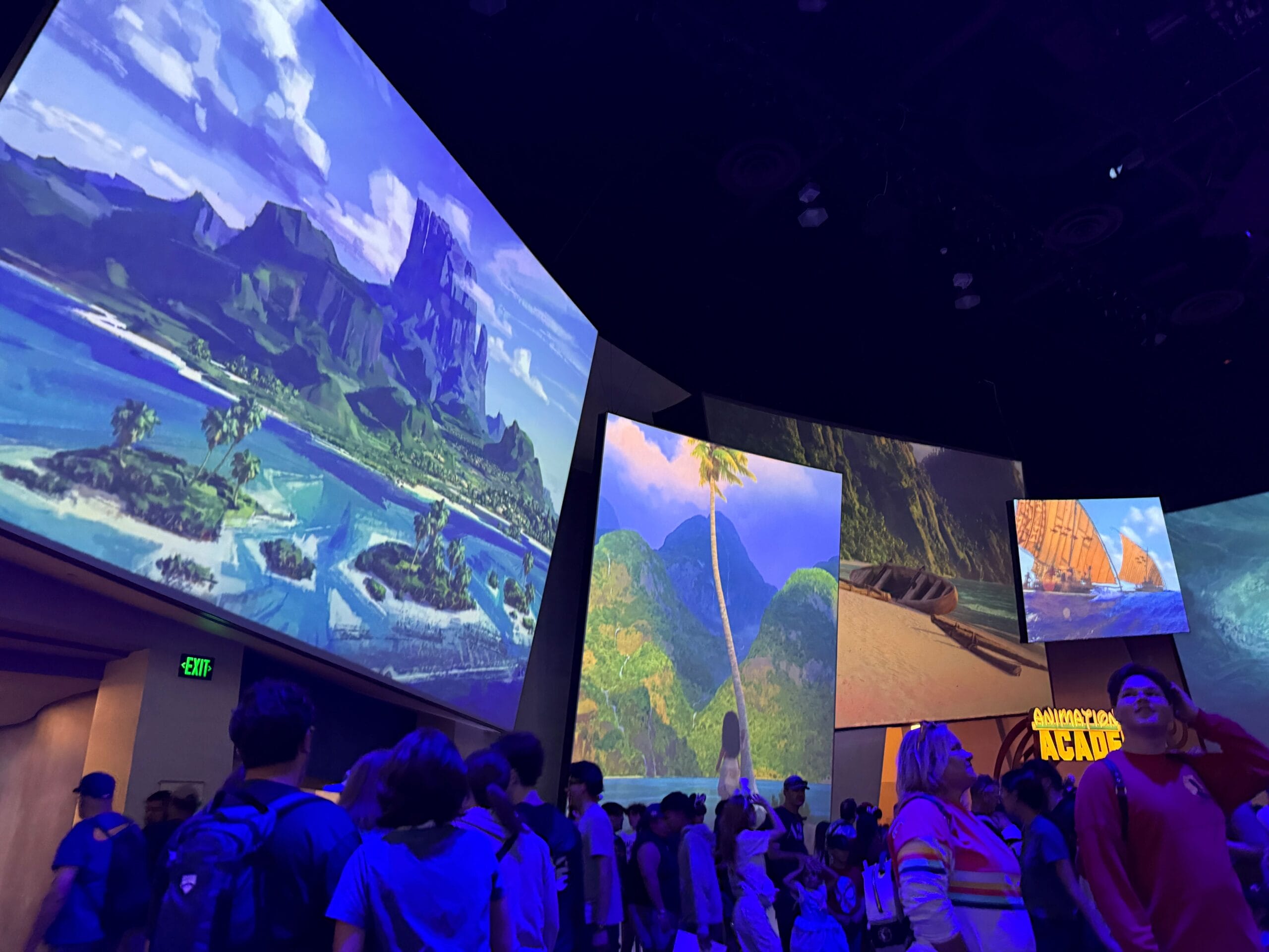 Crowd watching large screens displaying tropical landscapes and islands at an indoor event or exhibition.